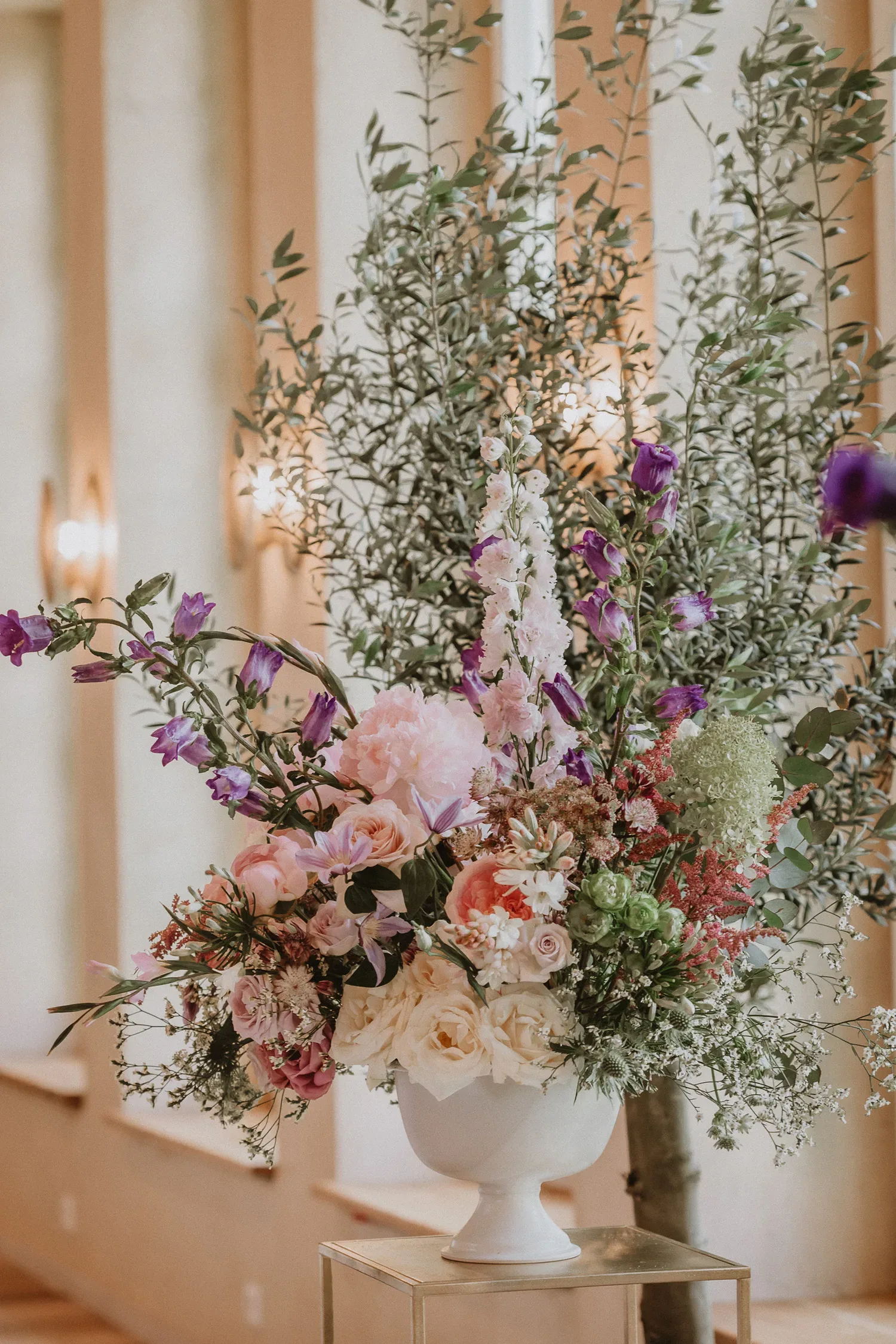 Ein großes Blumenarrangement in einer weißen Vase mit rosa Rosen, lila Blüten und grünen Akzenten, dekoriert vor einem eleganten Innenraum mit hohen Fenstern und Leuchten.