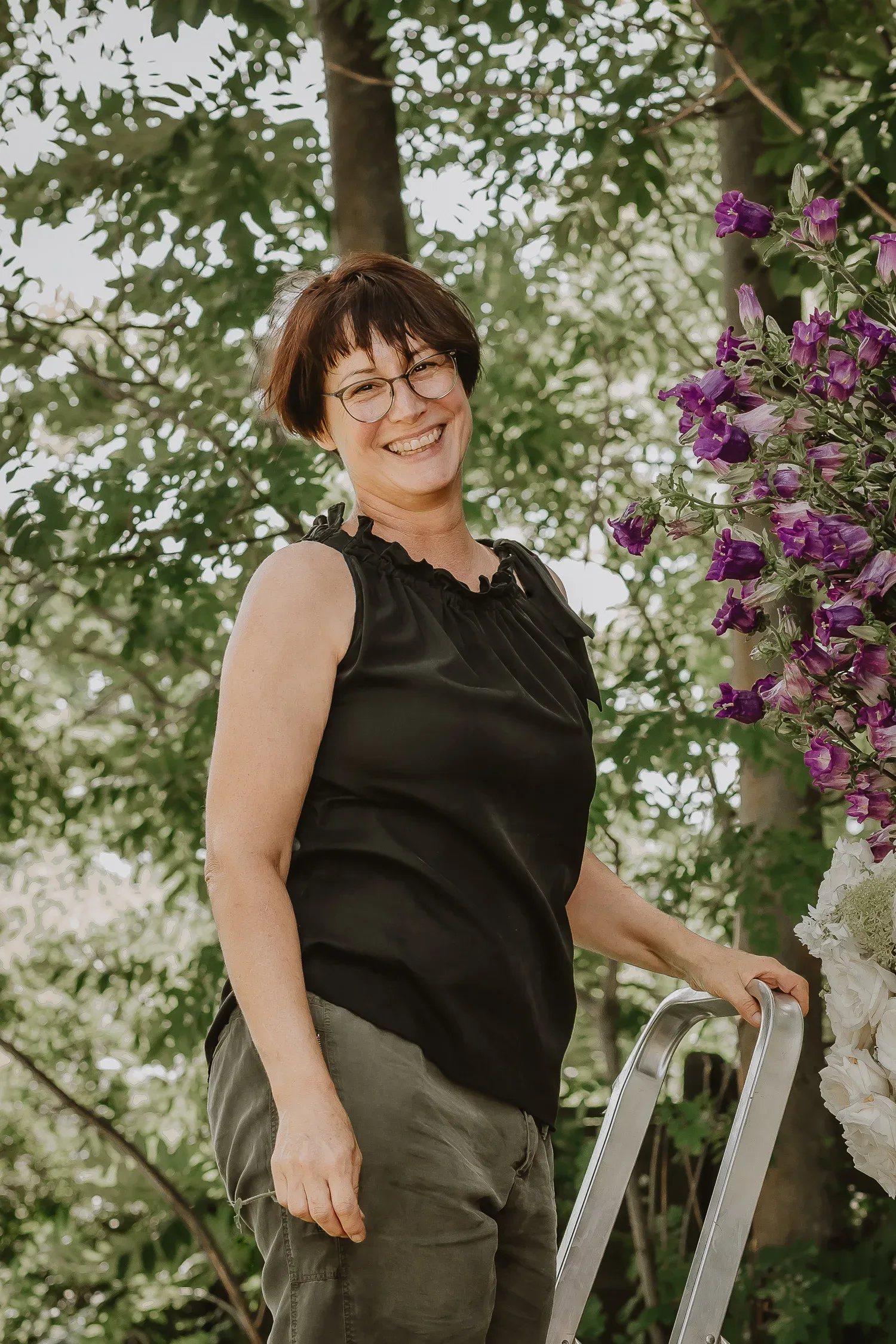 Eine Frau mit Brille und kurzem, braunem Haar lächelt freundlich in die Kamera, während sie neben einem üppigen Arrangement aus violetten Blumen steht.