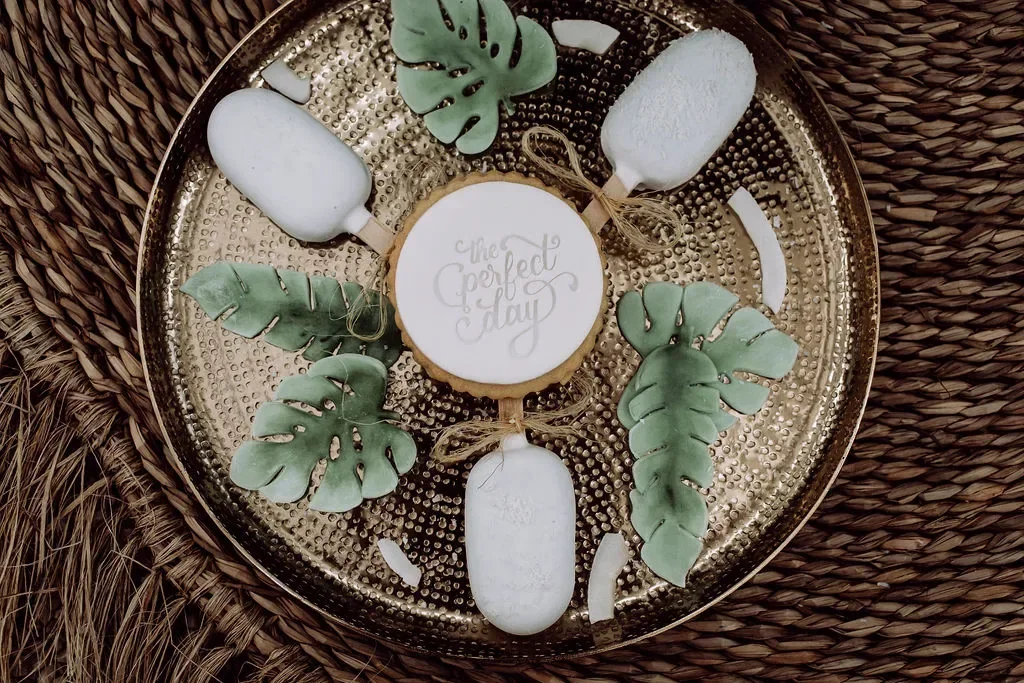 Tropische Hochzeitssüßigkeiten auf einem goldenen Tablett: dekorative Kekse und weiße Popsicles mit Monstera-Blättern.