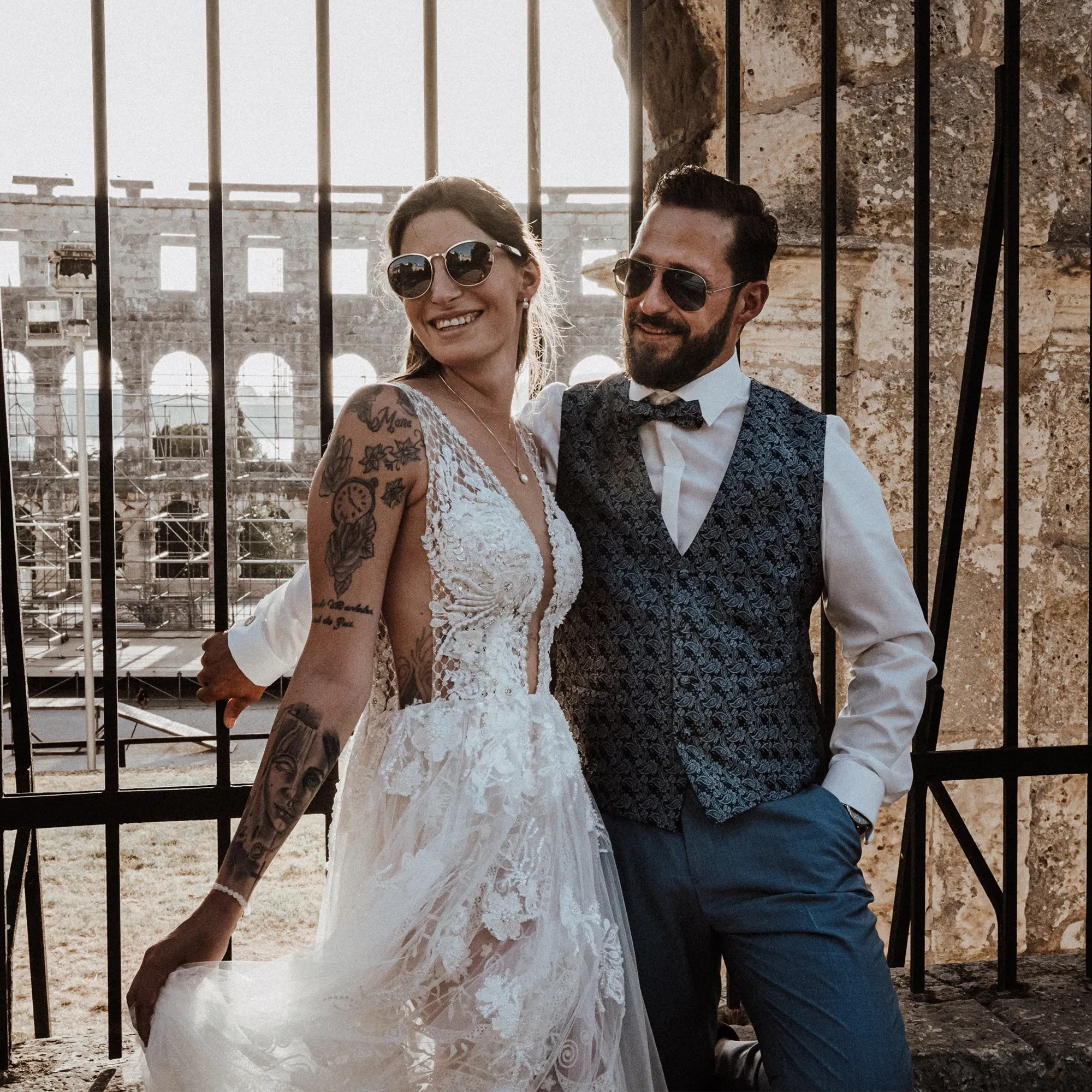 Braut und Bräutigam in stilvollen Sonnenbrillen posieren lächelnd in ihren Hochzeitsoutfits vor einem historischen Gebäude.