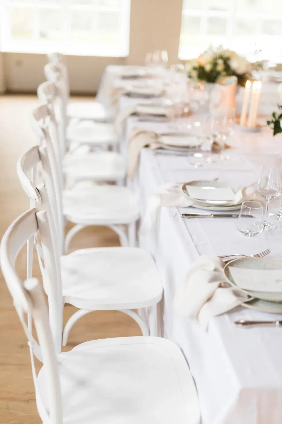 Weiß eingedeckte Hochzeitstafel mit stilvoll arrangierten Plätzen und minimalistischer Dekoration in einem hellen, luftigen Raum.