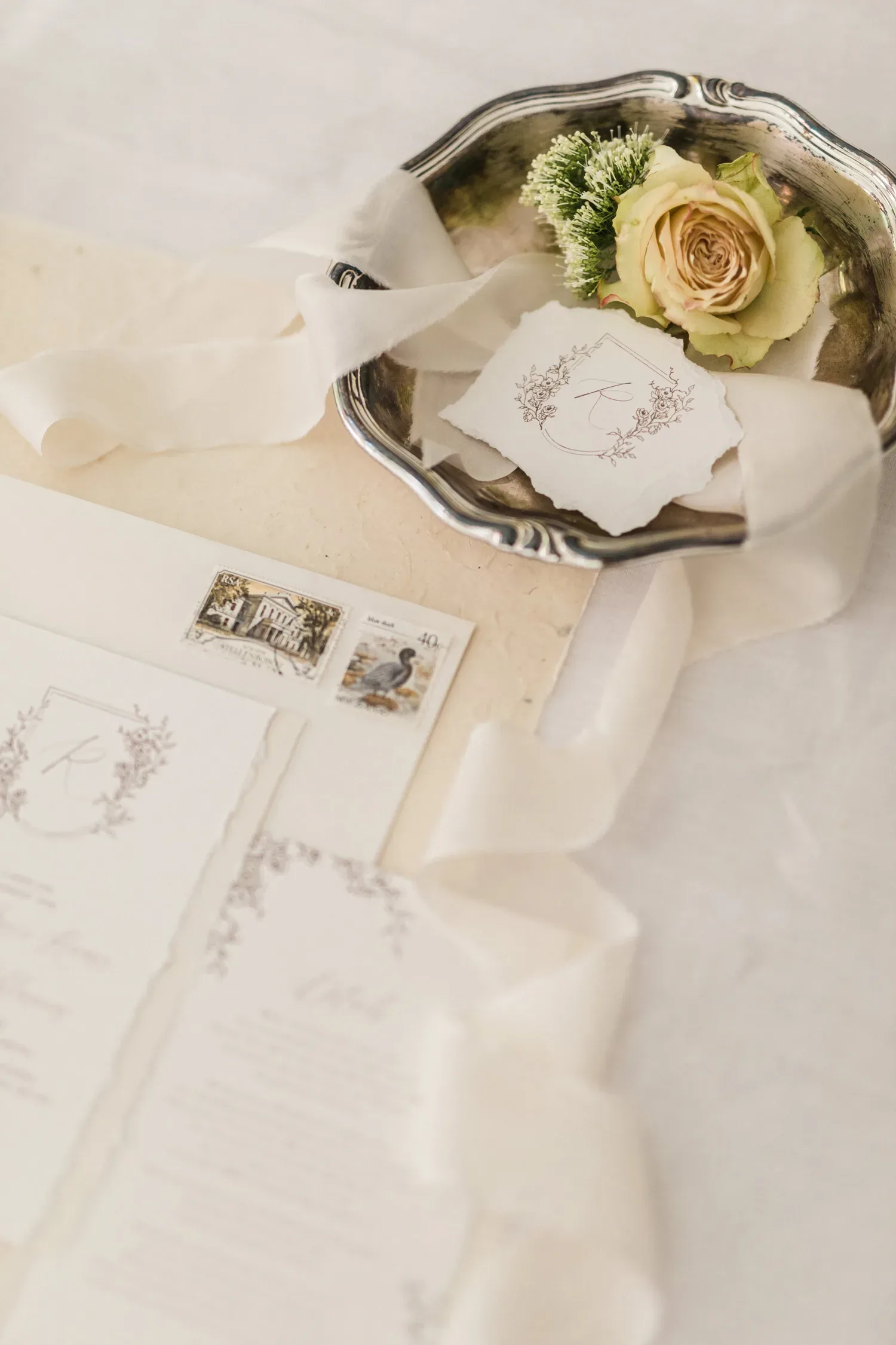 Eine silberne Schale mit einer zarten, cremefarbenen Rose und feiner Hochzeitspapeterie in einem eleganten, floralen Design.