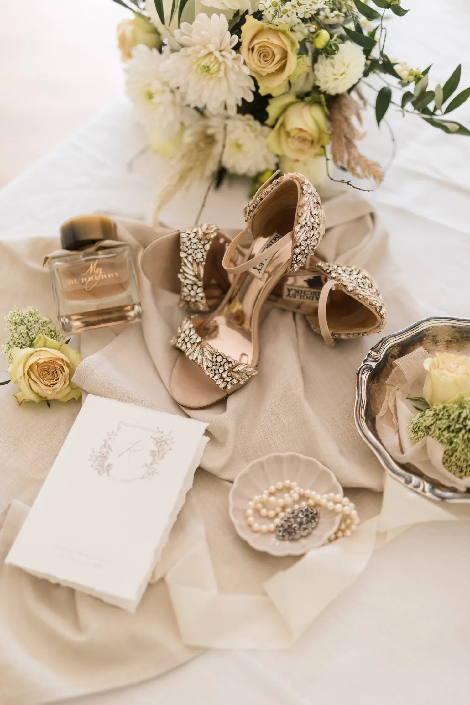 Auf einem sanft drapierten Leinentuch liegen sorgfältig arrangierte Hochzeitsaccessoires: glänzende Sandaletten mit Kristallapplikationen, eine stilvolle Parfümflasche, eine zarte Perlenkette in einer Schale sowie eine florale Hochzeitseinladung. Ein Bouquet aus cremefarbenen Blüten rundet das romantische Ensemble ab.