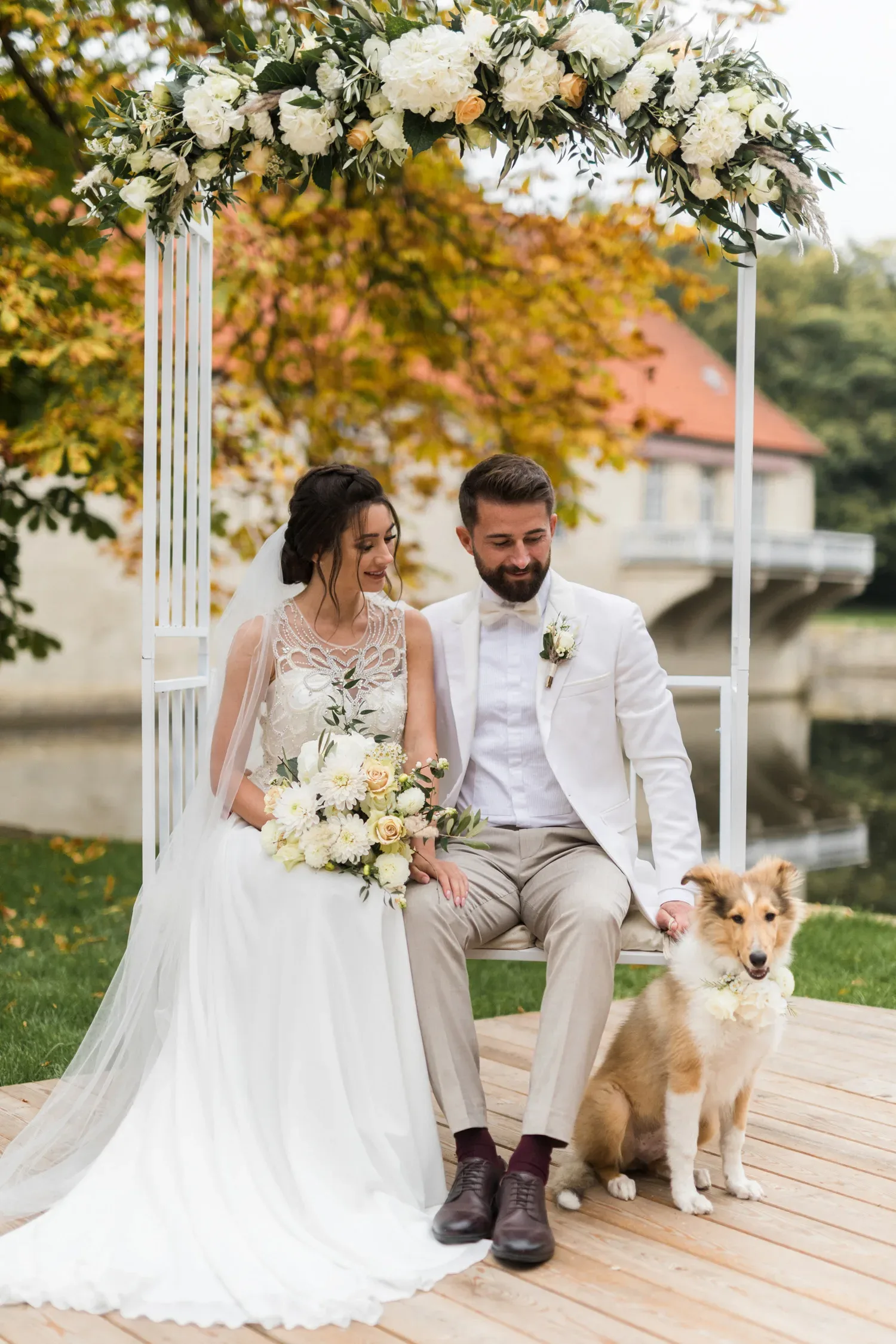 Das Brautpaar sitzt glücklich unter einem üppigen Blumenbogen, der mit weißen und cremefarbenen Blüten geschmückt ist. Sie lächeln, während ein niedlicher Hund mit einem floralen Halsband neben ihnen sitzt. Der Hintergrund zeigt eine herbstliche Landschaft mit einem Gebäude und einem Fluss.