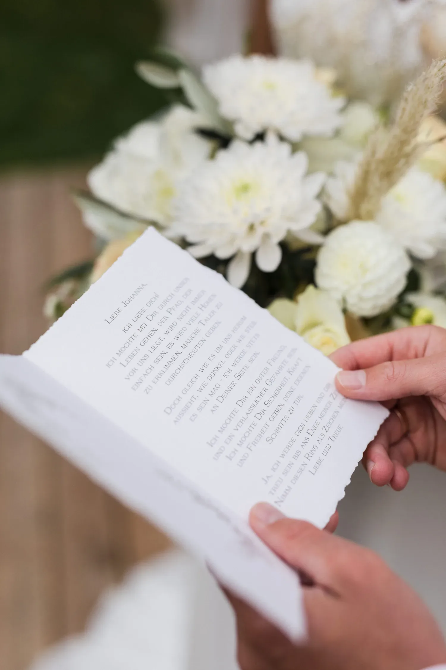 Nahaufnahme von zwei Händen, die ein handgeschriebenes Gelübde auf einem zart gerissenen Stück Papier halten. Im Hintergrund sind weiße Blumenarrangements zu sehen, die die feierliche und emotionale Atmosphäre unterstreichen.