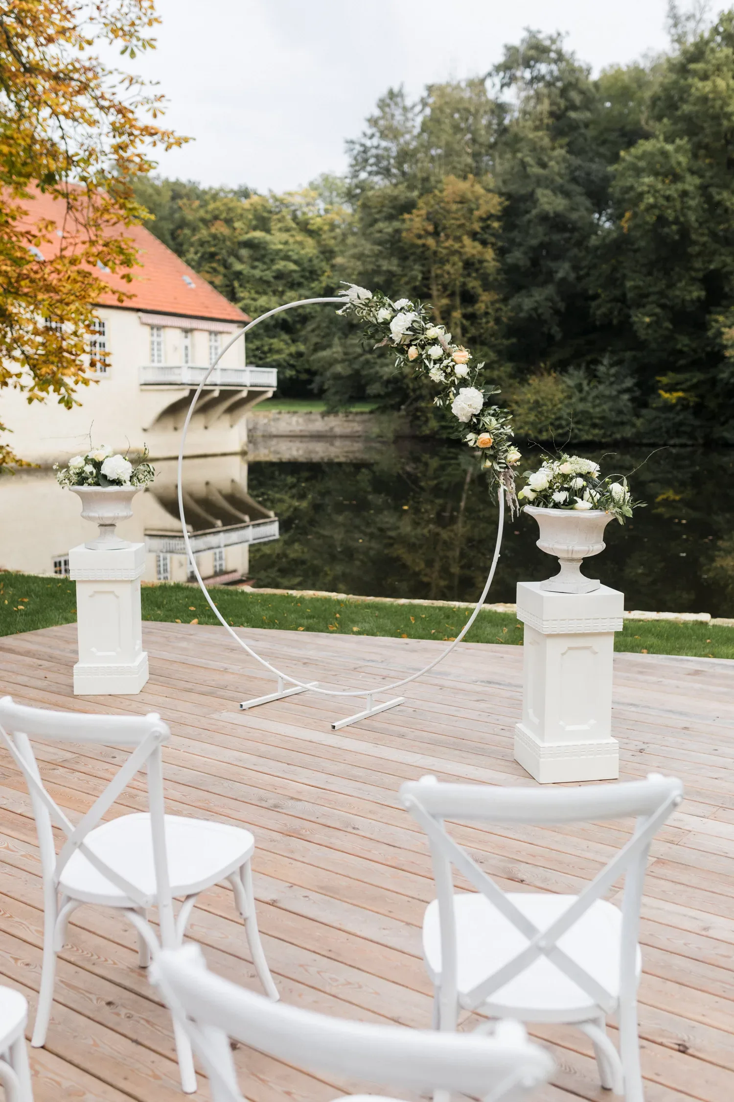 Eine schlichte und elegante Hochzeitszeremonie im Freien mit weißen Stühlen, einem runden Blumenbogen und einem malerischen Gebäude im Hintergrund, das sich im Wasser spiegelt. Der Altar ist mit weißen Blumen geschmückt.