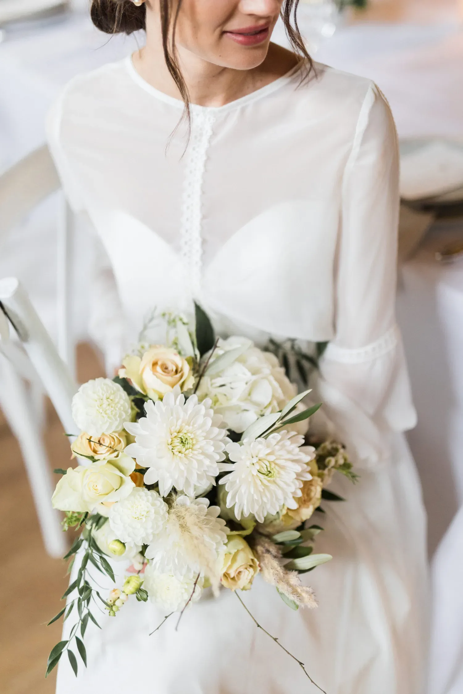 Eine Braut in einem schlichten, eleganten Kleid hält einen üppigen Blumenstrauß mit weißen Dahlien, cremefarbenen Rosen und grünen Blättern. Ihr zartes Lächeln und die dezente Hochzeitsdekoration im Hintergrund vervollständigen die Szene.