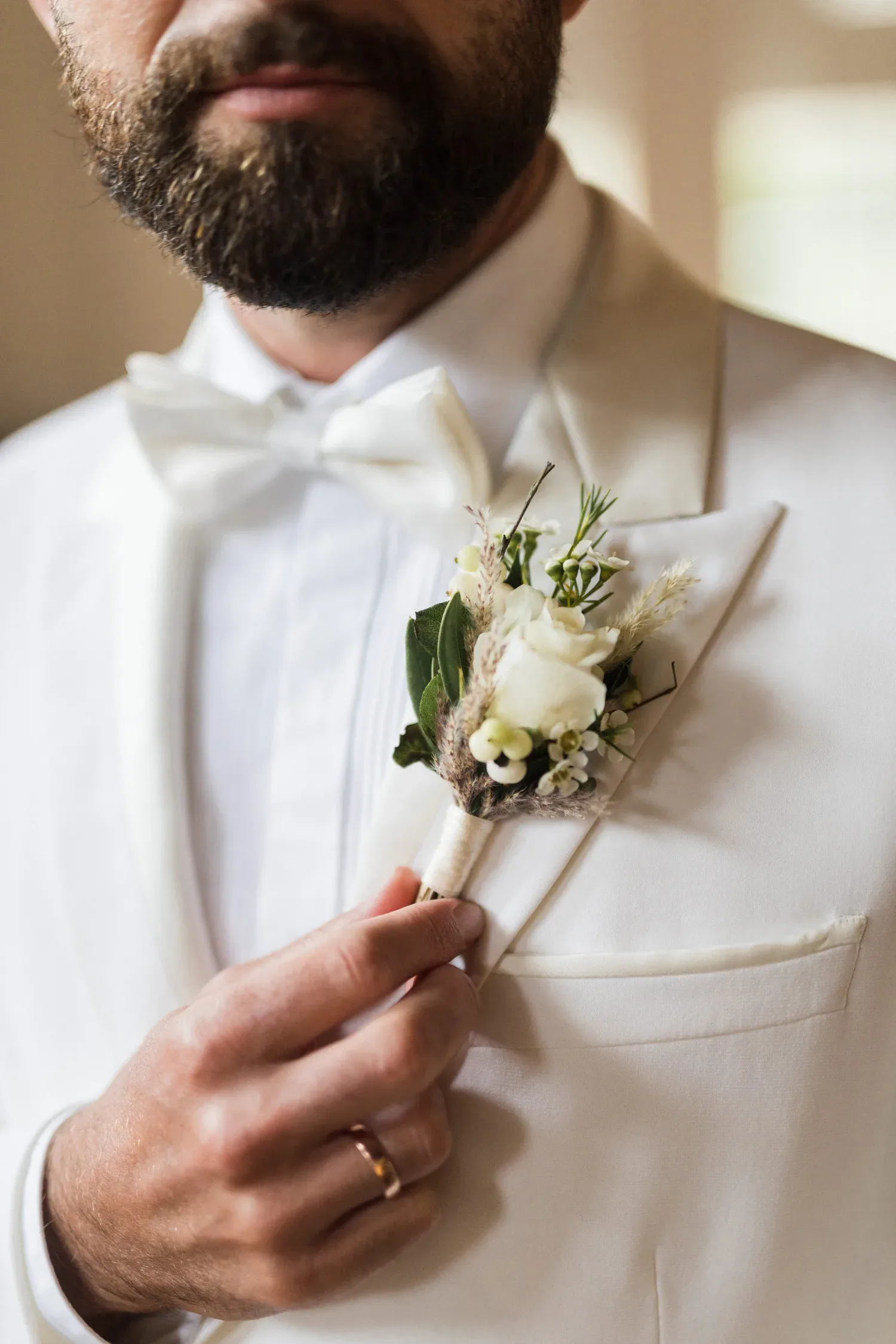 Ein Bräutigam in einem cremefarbenen Anzug richtet seine Blumenanstecknadel, die aus weißen Rosen und kleinen grünen Akzenten besteht. Die dezente Eleganz seines Outfits wird durch eine weiße Fliege ergänzt.