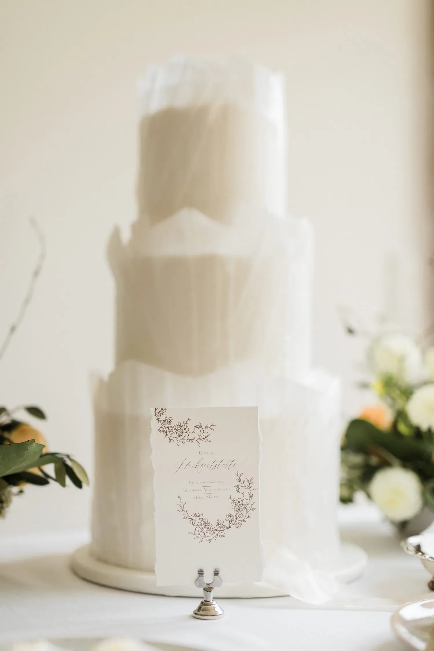 Eine elegante Hochzeitstorte mit mehreren Etagen, umhüllt von zarter, weißer Fondantverzierung. Im Vordergrund steht eine handgeschriebene Menükarte, die auf einem kleinen silbernen Kartenhalter präsentiert wird. Blumenarrangements in Weiß und Grün zieren den Tisch.
