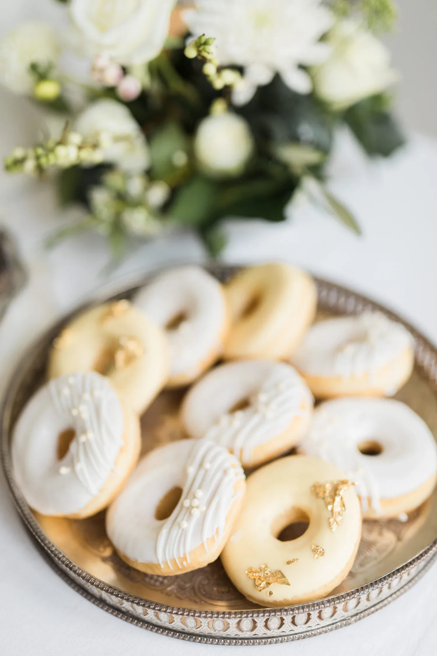 Ein silbernes Tablett, dekoriert mit cremeweißen Donuts, die mit feinen Perlen und goldenen Akzenten verziert sind. Im Hintergrund ist ein Blumenarrangement in Weiß und Grün sichtbar, das den festlichen Tisch schmückt.