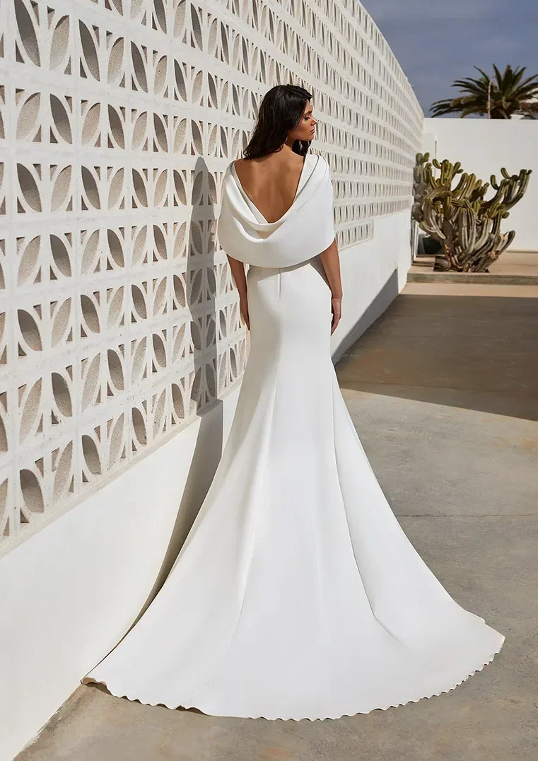 Rückansicht eines eleganten, weißen Brautkleids mit drapiertem Rückenausschnitt und langer Schleppe, getragen von einem Modell, das an einer gemusterten weißen Wand entlanggeht.