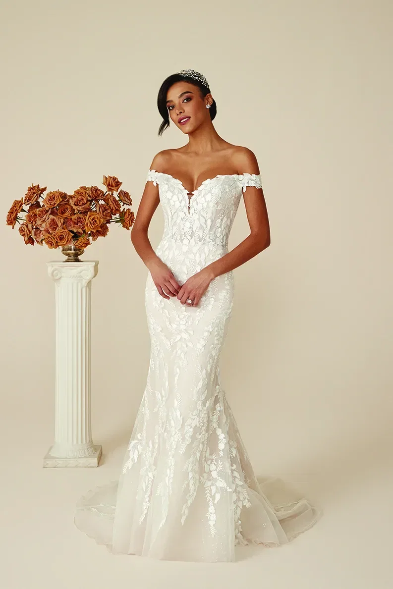 Braut trägt ein schulterfreies, figurnahes Brautkleid mit floralen Stickereien, posiert neben einer Säule mit Trockenblumen, in einem eleganten Studio-Setting.