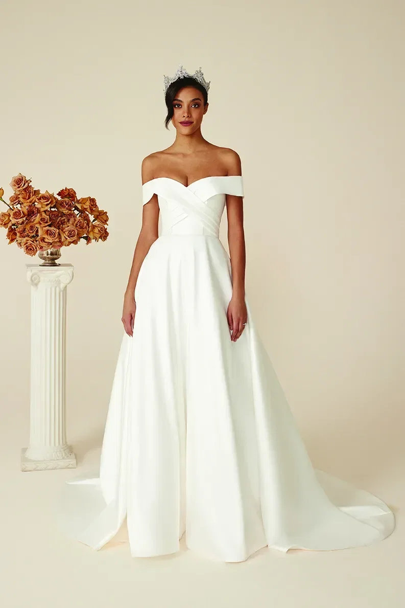 Brautmodell trägt ein schlichtes, schulterfreies Ballkleid mit herzförmigem Ausschnitt und Krone, posiert neben einer Säule mit herbstlichen Blumenarrangements.