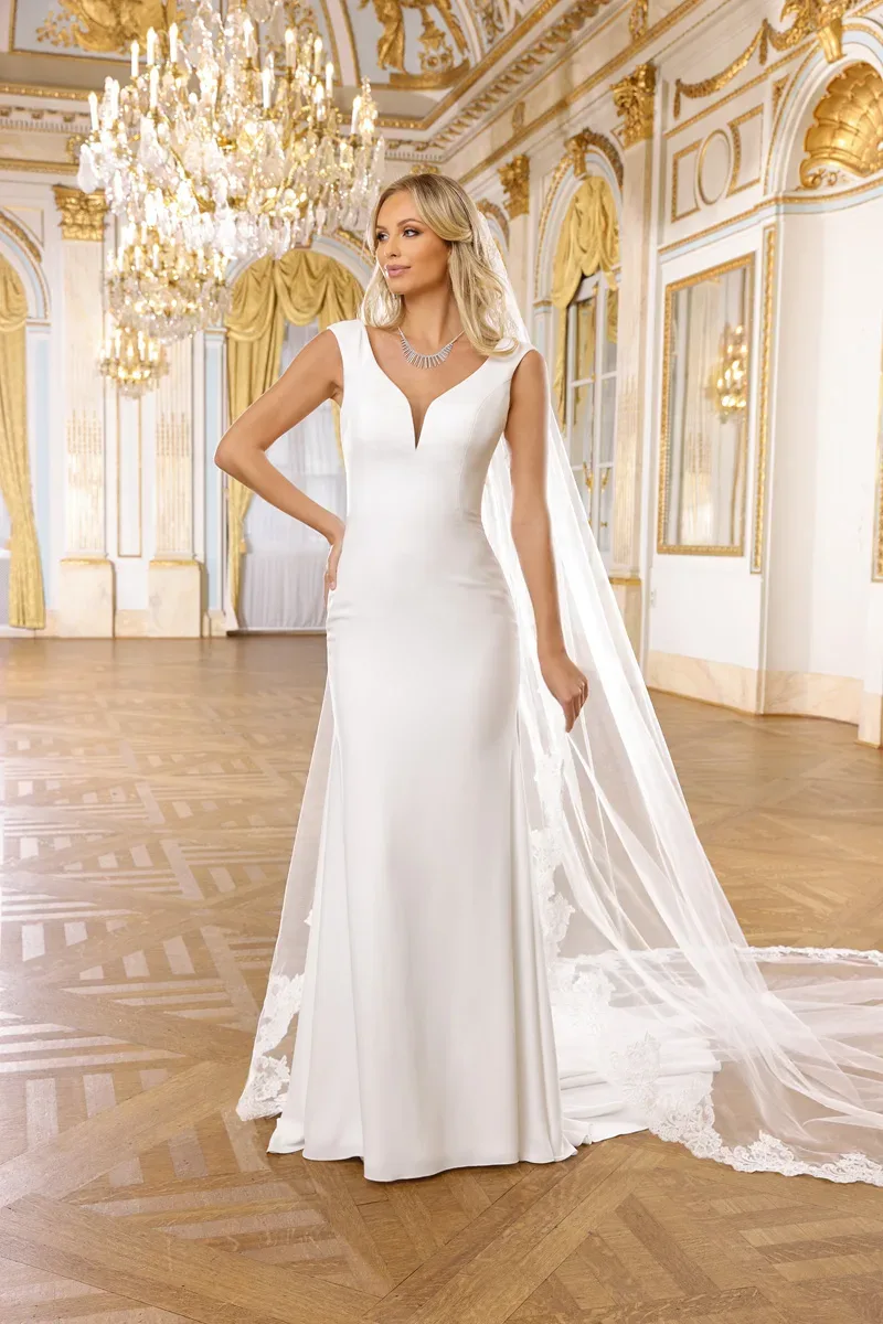 Braut in einem eleganten, ärmellosen Brautkleid mit schlichtem Design und langer Schleppe posiert in einem prunkvollen Raum mit Kristallleuchtern und aufwendig dekorierten Wänden.