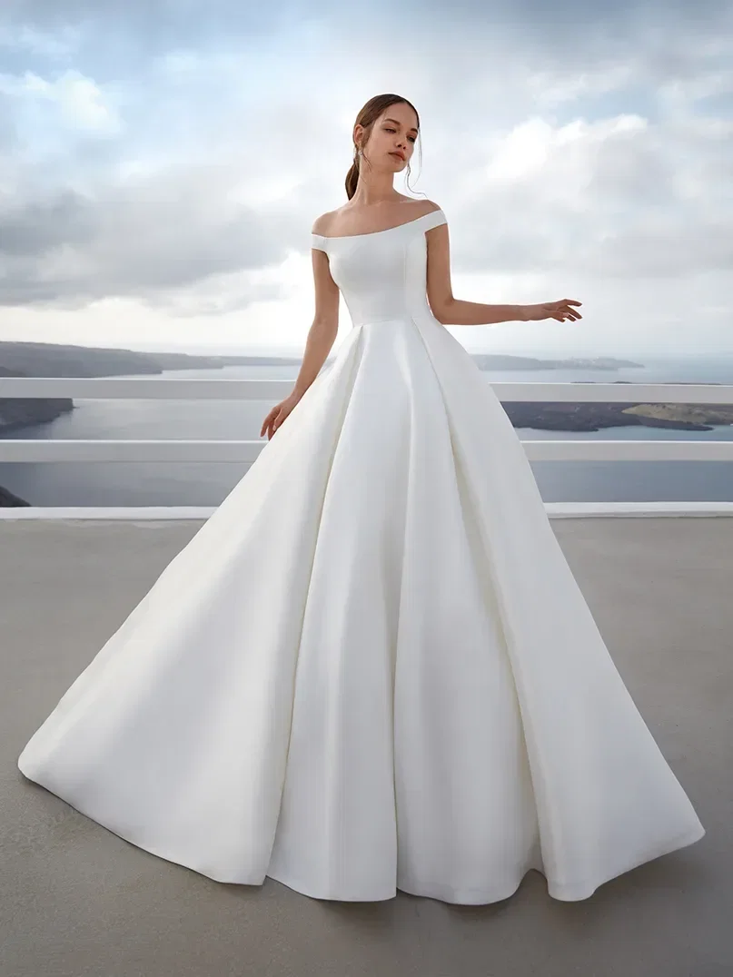 Braut in einem klassischen, weißen, schulterfreien Ballkleid mit voluminösem Rock, posiert vor einer ruhigen Küstenlandschaft mit weitem Blick über das Meer.