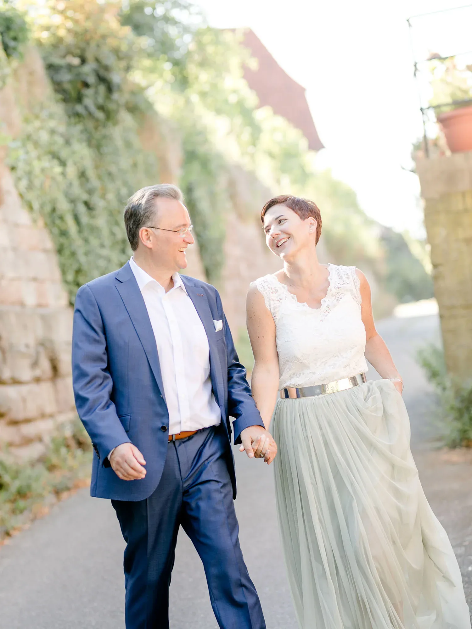 Ein lächelndes Paar, das Hand in Hand einen sonnigen Weg entlang spaziert. Der Mann trägt einen blauen Anzug, während die Frau ein cremefarbenes Oberteil und einen fließenden, hellen Rock trägt. Die Umgebung ist grün und von Mauern gesäumt.