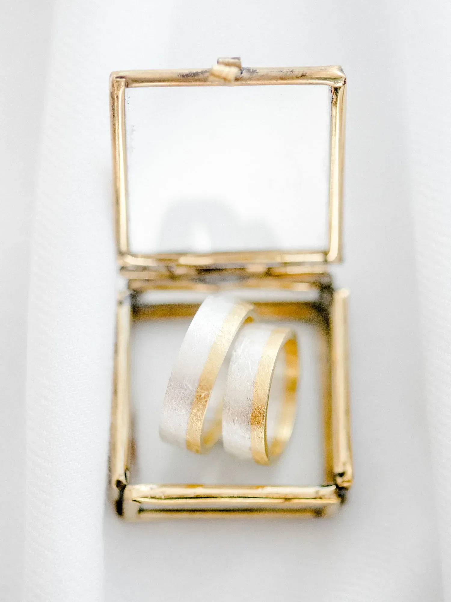 Zwei goldene Eheringe mit einem feinen, silbernen Streifen in einer eleganten, offenen Schmuckschatulle präsentiert. Die Schatulle ist aus goldenem Metall, und die Ringe liegen auf einem weißen Stoff.