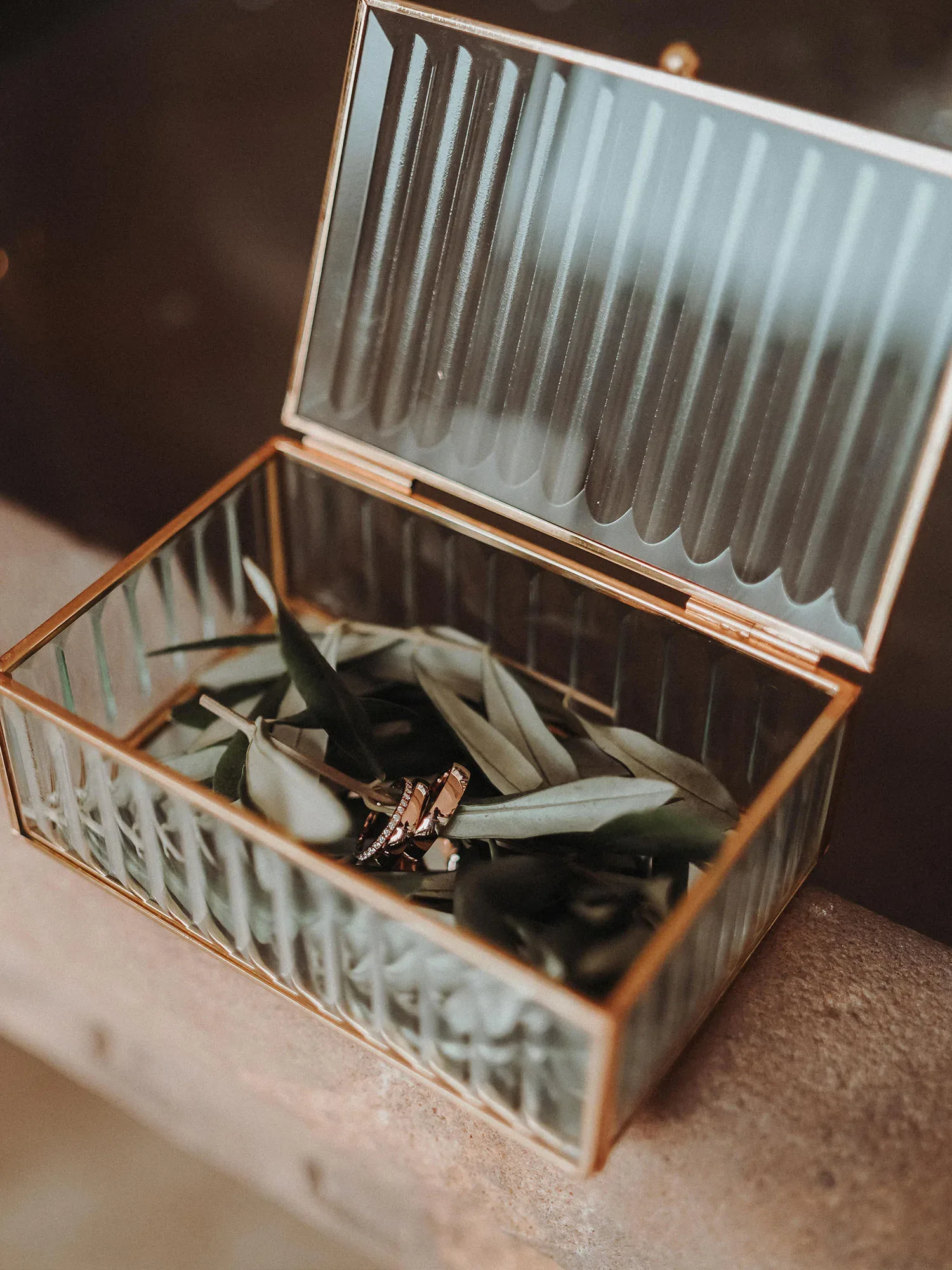 Eine geöffnete Glasbox, gefüllt mit Olivenblättern und Eheringen, steht auf einer rustikalen Steinoberfläche. Die Box ist mit einem goldenen Rahmen verziert und lässt den edlen Inhalt elegant zur Geltung kommen.