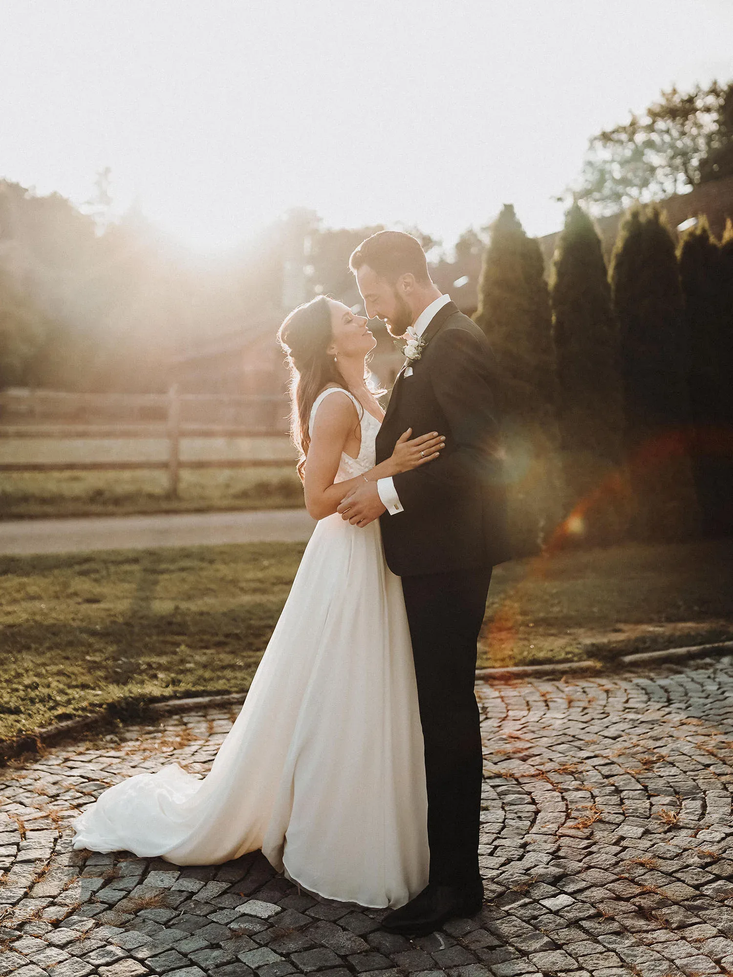 Ein Hochzeitspaar tanzt in der Abendsonne auf einem gepflasterten Platz. Die Braut trägt ein weißes, fließendes Kleid, während der Bräutigam einen eleganten schwarzen Anzug trägt. Das Paar schaut sich verliebt an, während das Licht der untergehenden Sonne die Szene sanft beleuchtet.
