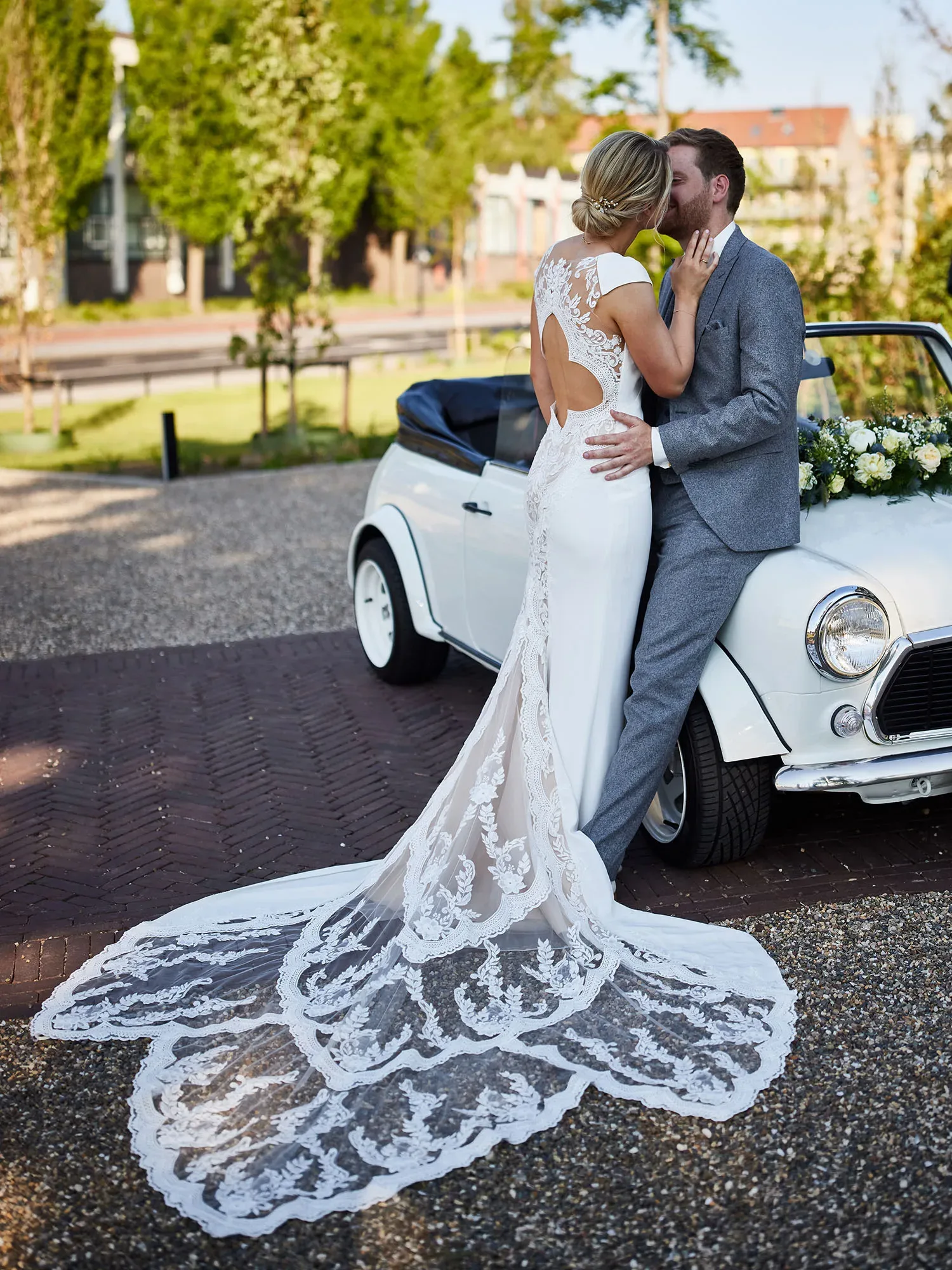 Ein Brautpaar küsst sich vor einem weißen, vintage Cabrio. Die Braut trägt ein Kleid mit einer langen, bestickten Schleppe, während der Bräutigam im grauen Anzug sich gegen das Auto lehnt. Die Szene findet im Freien bei sonnigem Wetter statt.