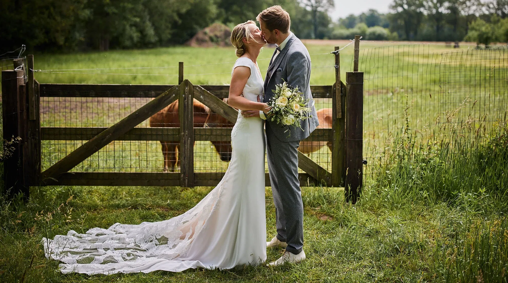 Ein Brautpaar küsst sich vor einem Holztor auf einem ländlichen Feld. Die Braut trägt ein schlichtes, elegantes Kleid mit langer Schleppe, und der Bräutigam trägt einen grauen Anzug. Im Hintergrund sind Kühe auf der Weide zu sehen.