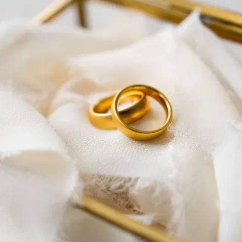 Zwei schlichte, goldene Eheringe liegen auf einem weichen, cremefarbenen Stoff in einer eleganten Glasschatulle, bereit für die Trauzeremonie.