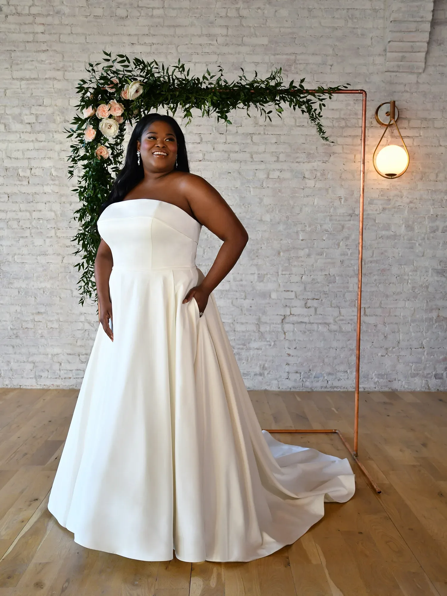 Eine Braut in einem trägerlosen, eleganten Hochzeitskleid mit A-Linie und Schleppe lächelt auf einem Holzfußboden vor einem Blumenbogen mit Rosen und Eukalyptusblättern.