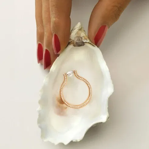 Eine Hand mit roten, lackierten Nägeln hält eine offene Muschelschale, in der ein zarter, roségoldener Verlobungsring liegt. Der Ring schimmert auf der weißen Innenseite der Muschel.