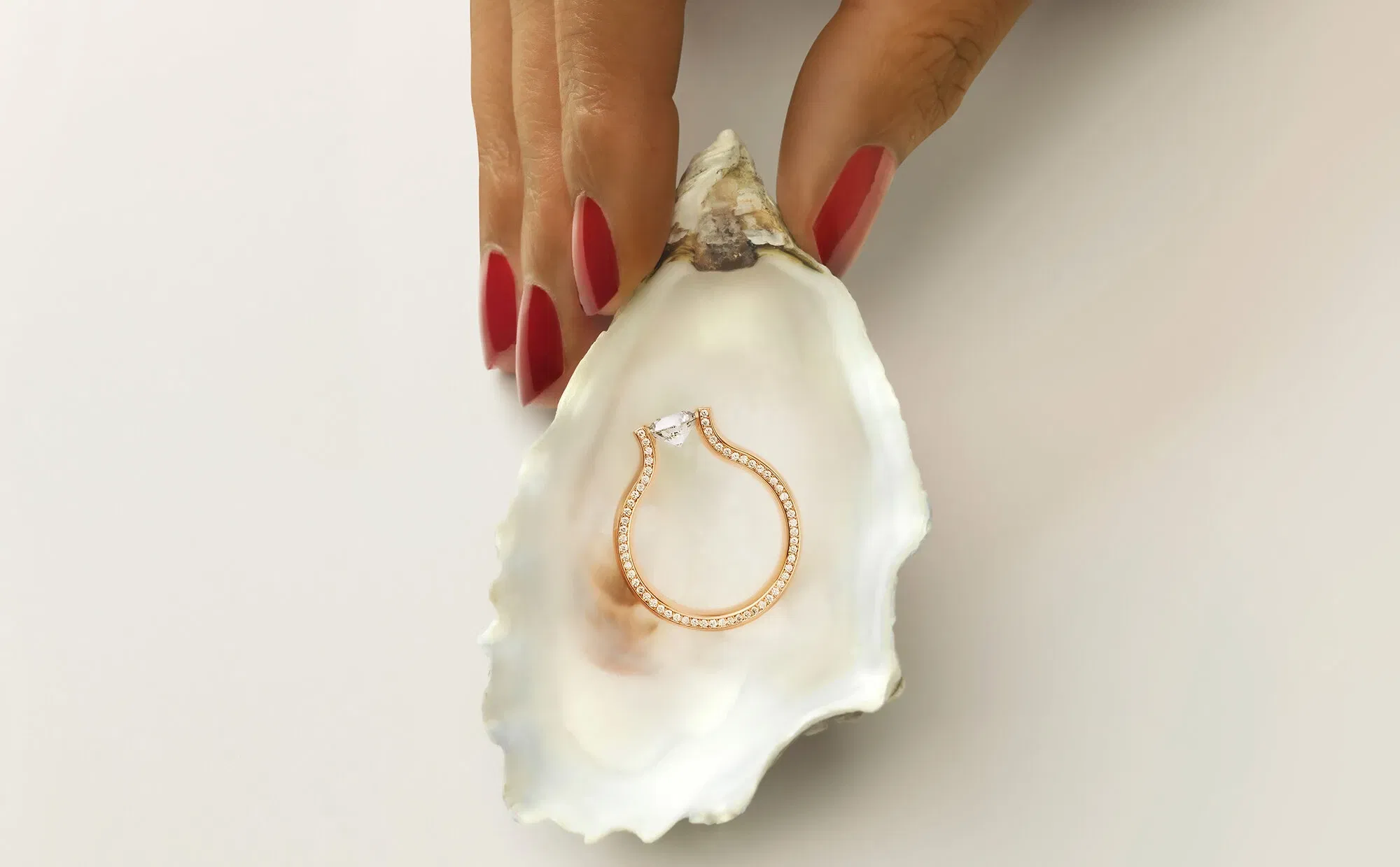 Eine Hand mit roten, lackierten Nägeln hält eine offene Muschelschale, in der ein zarter, roségoldener Verlobungsring liegt. Der Ring schimmert auf der weißen Innenseite der Muschel.