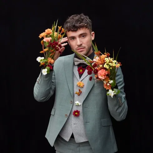 Ein Mann in einem pastellgrünen Anzug posiert vor einem schwarzen Hintergrund, während er bunte Blumenarrangements in den Händen und am Anzug trägt.