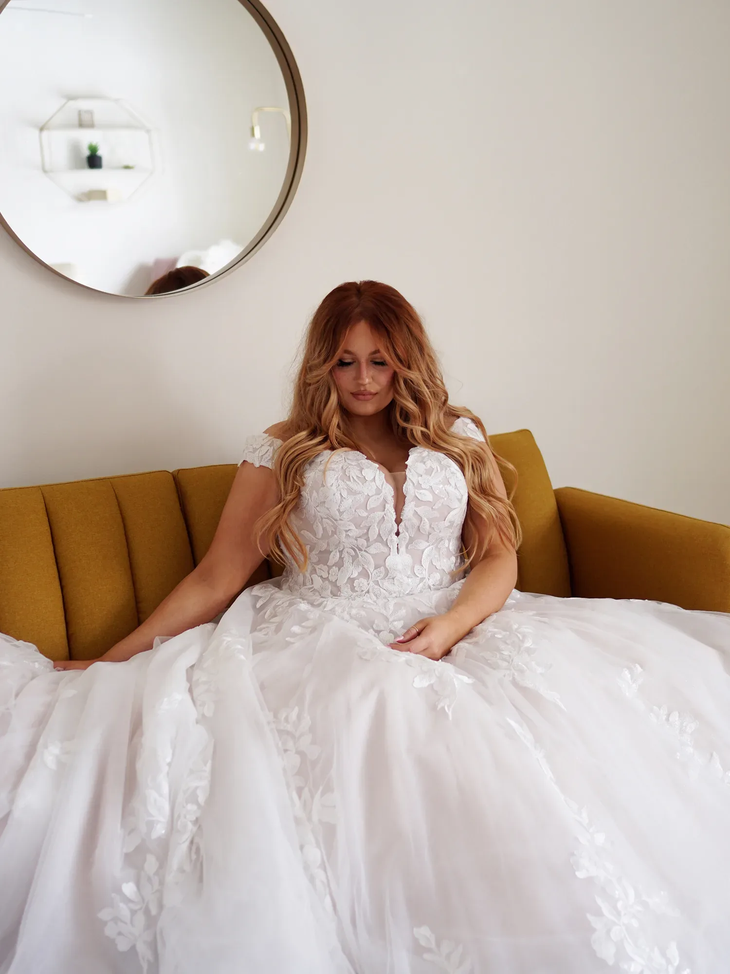 Braut sitzt entspannt auf einem gelben Sofa und trägt ein aufwendiges, weißes Brautkleid mit floralen Spitzenapplikationen und tiefem Ausschnitt, im Hintergrund ein runder Spiegel an der Wand.