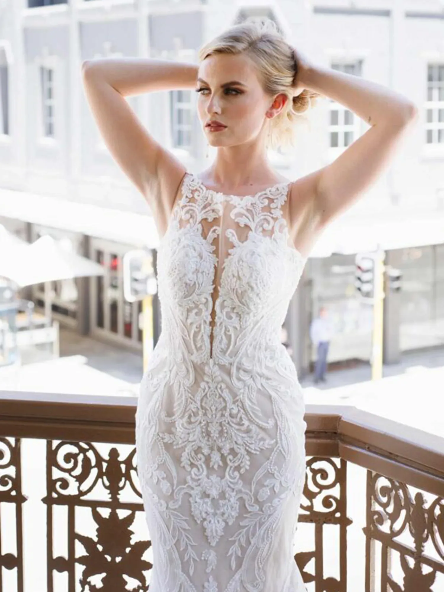 Eine Braut in einem eleganten Hochzeitskleid mit transparentem Oberteil und filigranen Stickereien steht auf einem Balkon und hebt die Arme über den Kopf, während sie entschlossen in die Ferne blickt.