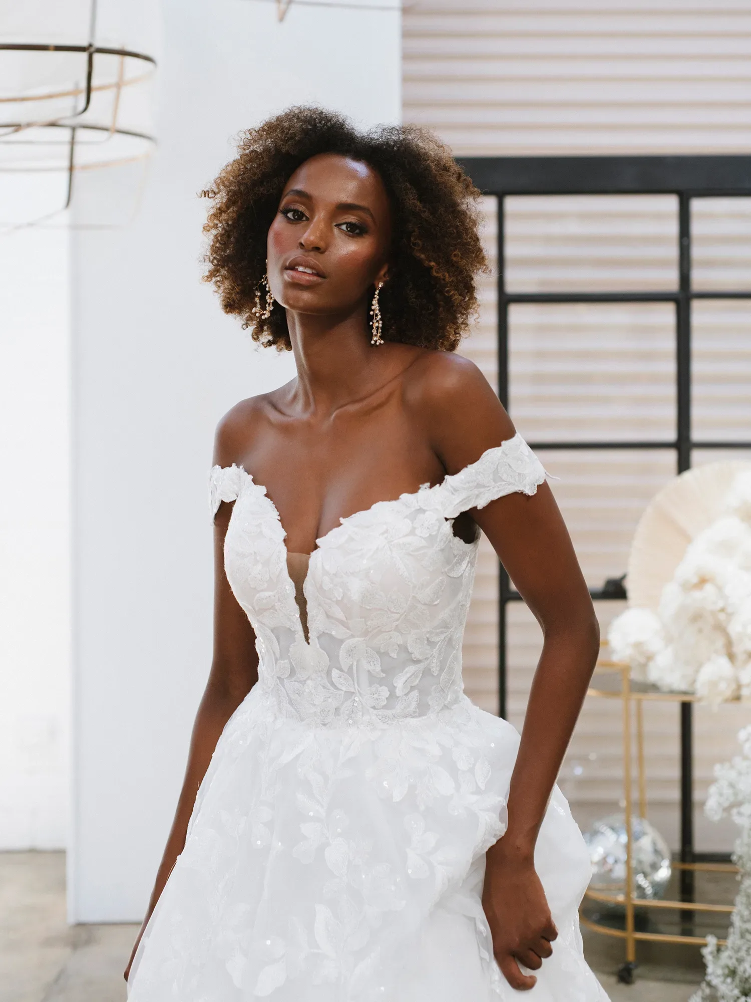 Nahaufnahme einer Braut in einem schulterfreien Brautkleid mit tiefem Ausschnitt und floralen Spitzenapplikationen, das Modell steht in einem modernen Raum und trägt auffällige Ohrringe.