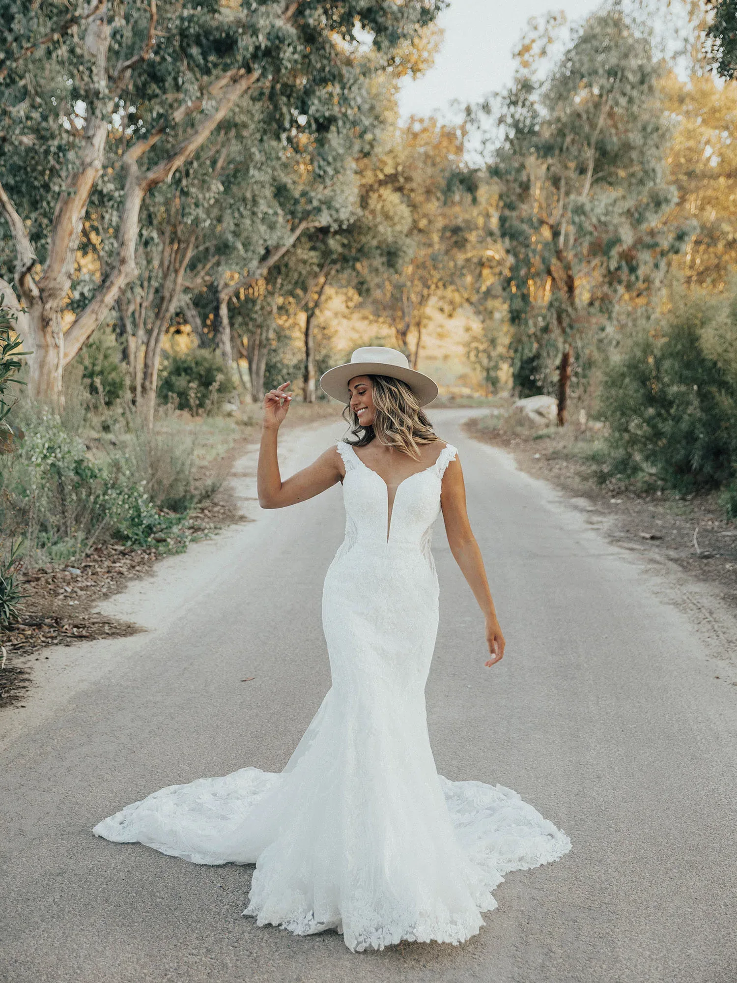 "Eine Braut in einem Spitzen-Hochzeitskleid mit tiefem Ausschnitt und Schleppe spaziert fröhlich auf einer ländlichen Straße, während sie einen weißen Hut trägt und von Bäumen umgeben ist.