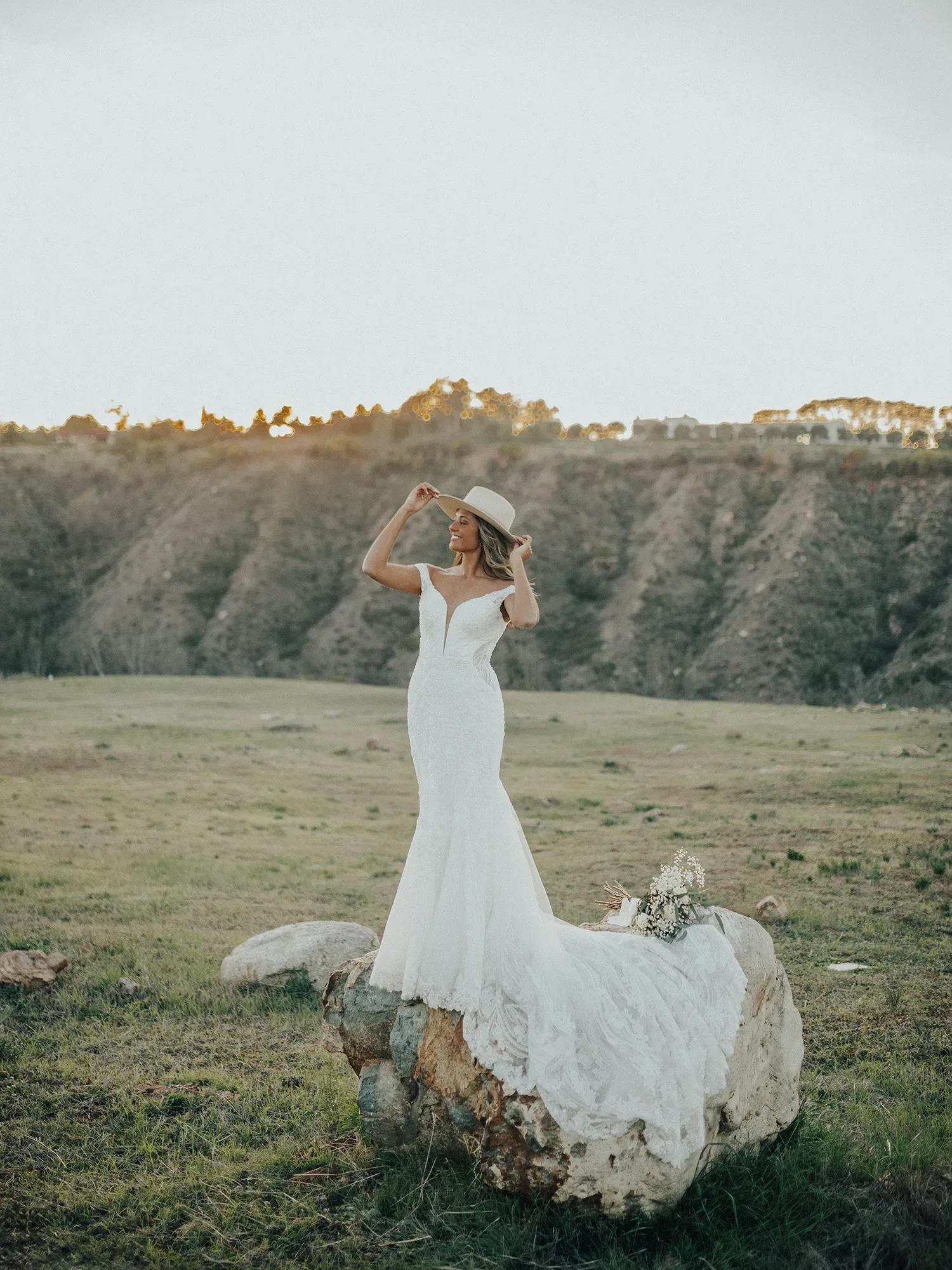 Eine Braut in einem Spitzen-Hochzeitskleid steht auf einem Felsen in einer weitläufigen Landschaft bei Sonnenuntergang, während sie ihren Hut leicht anhebt und in die Ferne blickt.