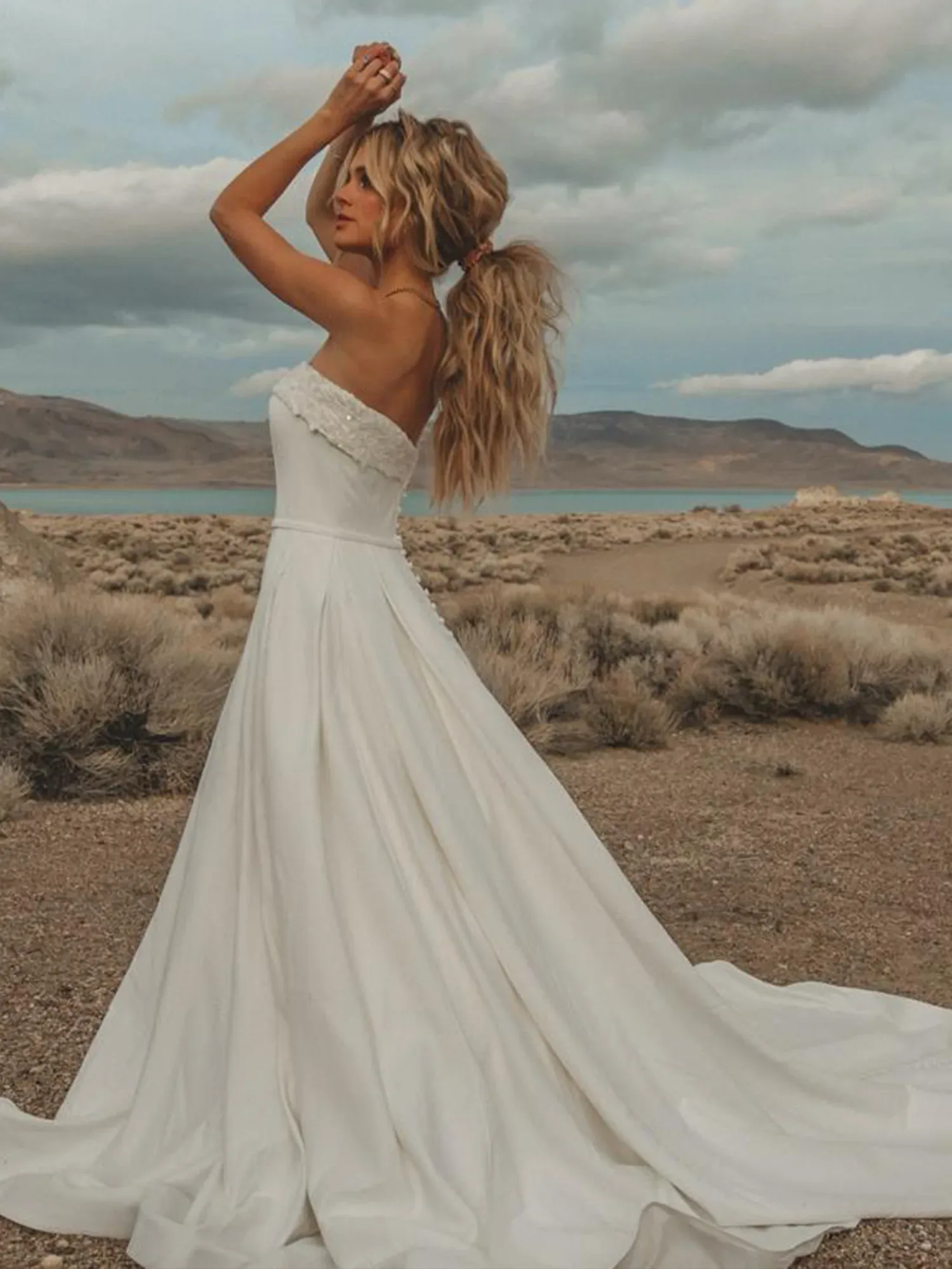 Eine Braut in einem trägerlosen Hochzeitskleid mit fließendem Rock und Schleppe posiert in einer Wüstenlandschaft, während sie ihre Arme nach oben streckt und im Wind weht.