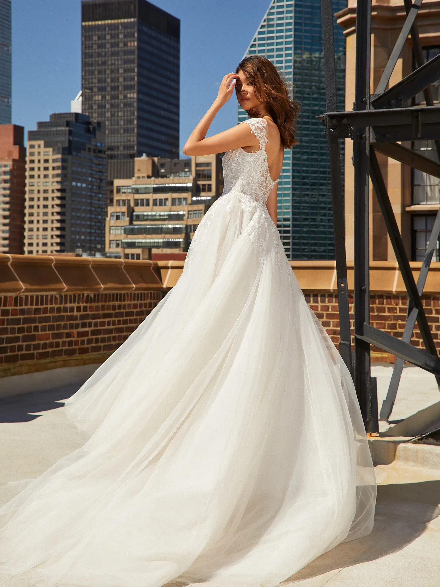 Eine Braut steht auf einer städtischen Dachterrasse und blickt über die Schulter zurück. Sie trägt ein atemberaubendes Kleid mit Spitzenrücken und einem weiten, fließenden Tüllrock, der elegant im Wind weht, während im Hintergrund moderne Wolkenkratzer die Skyline dominieren.