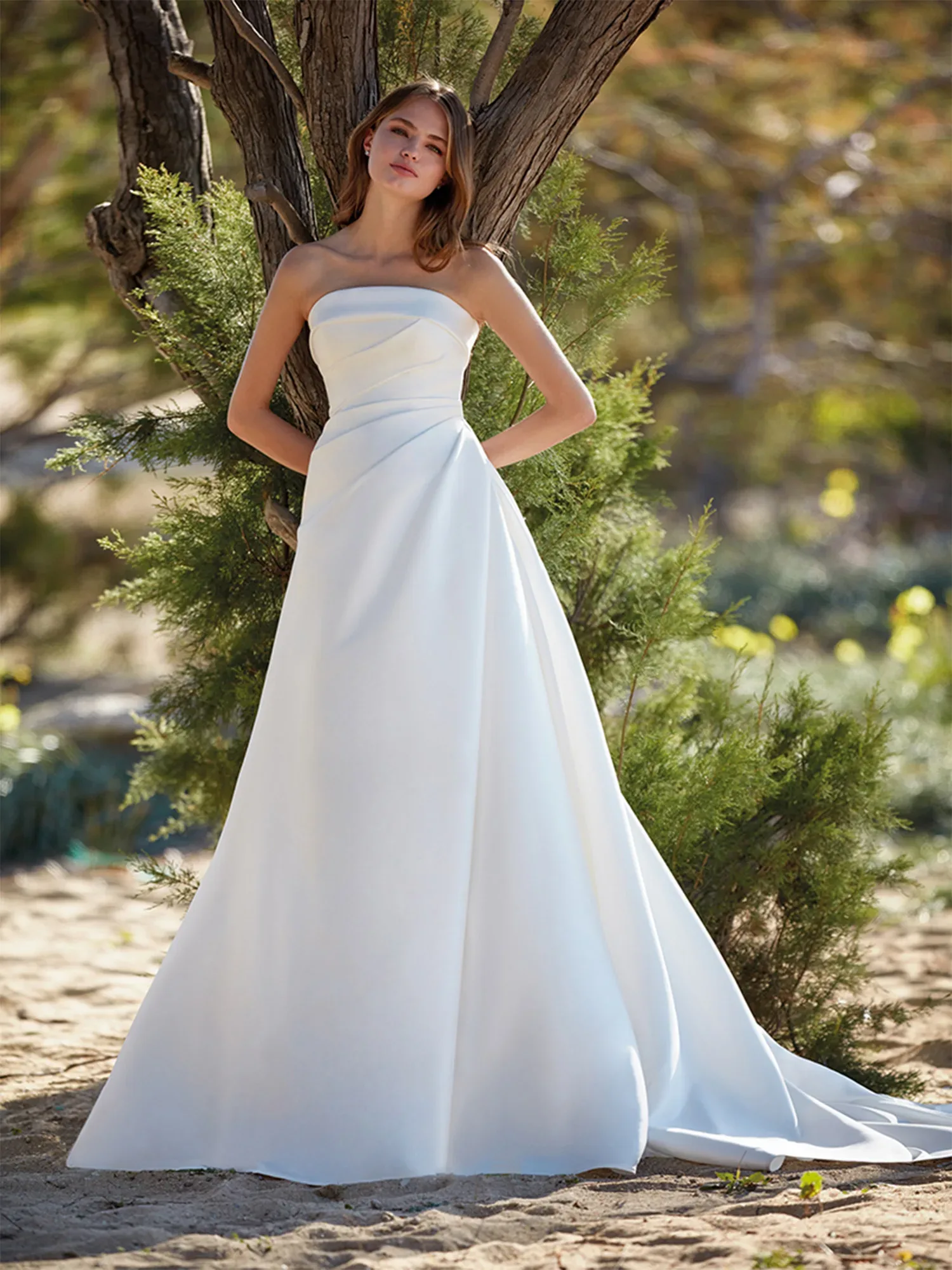 Eine junge Braut in einem schlichten, trägerlosen Brautkleid lehnt an einem Baum. Das fließende Kleid betont ihre elegante Figur, während die natürliche Umgebung im Hintergrund durch grünes Laub und sandigen Boden hervorgehoben wird.