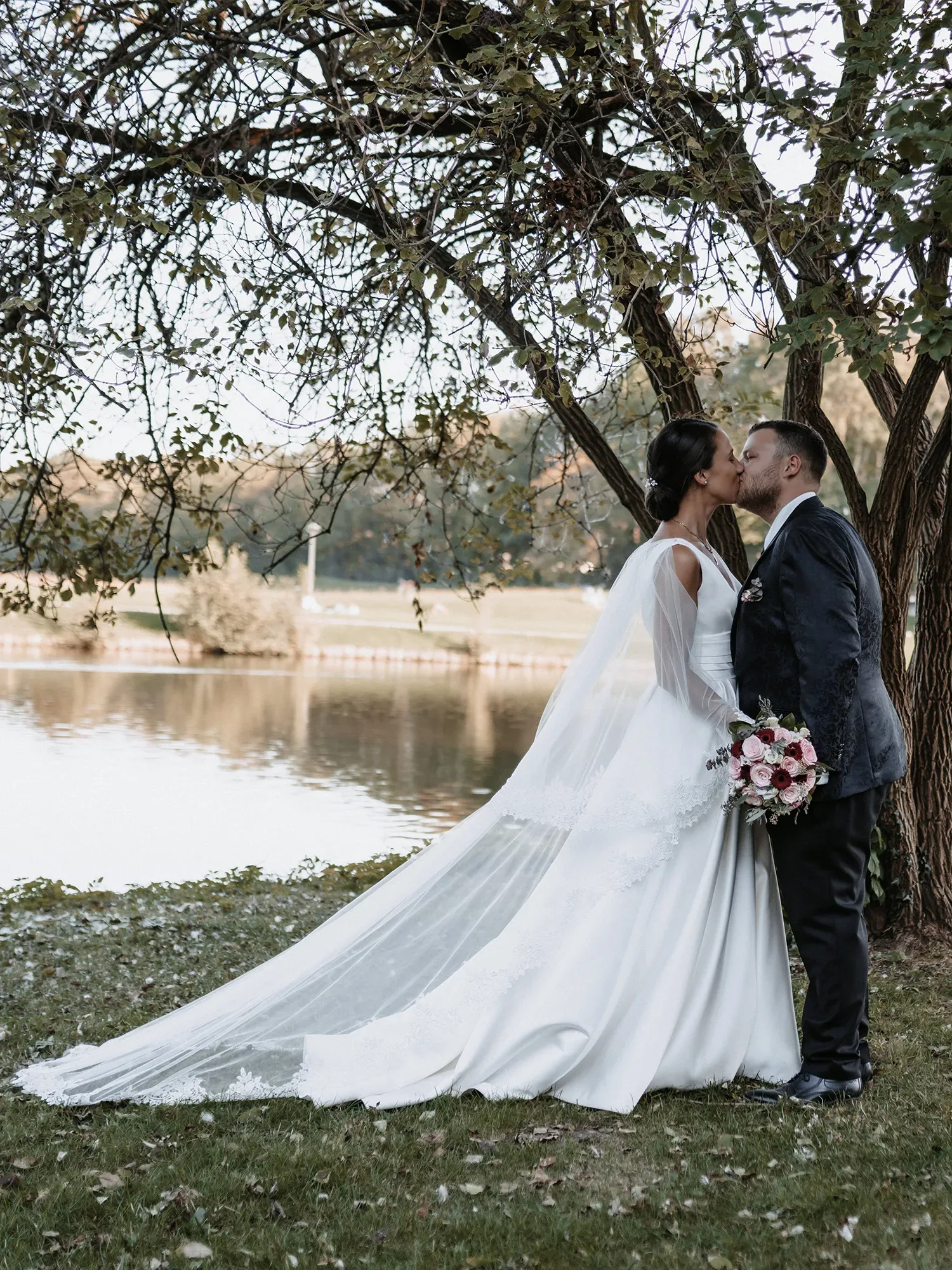 Claudia und Hendrik küssen sich unter einem Baum am Ufer eines ruhigen Sees. Die Braut trägt ein langes, fließendes Hochzeitskleid mit einer eleganten Schleppe und einem Schleier, während der Bräutigam einen dunklen Anzug trägt. Die natürliche Umgebung und das reflektierende Wasser schaffen eine romantische Atmosphäre.