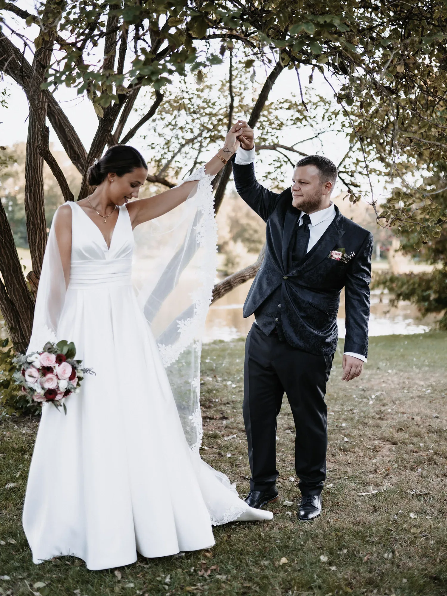 Claudia und Hendrik stehen unter einem Baum am Seeufer und halten sich an den Händen. Die Braut trägt ein schlichtes, elegantes weißes Hochzeitskleid mit einem langen Schleier, während der Bräutigam einen schwarzen Anzug trägt. Claudia hält einen Strauß rosafarbener Blumen, und die Szene strahlt Ruhe und Zuneigung aus.