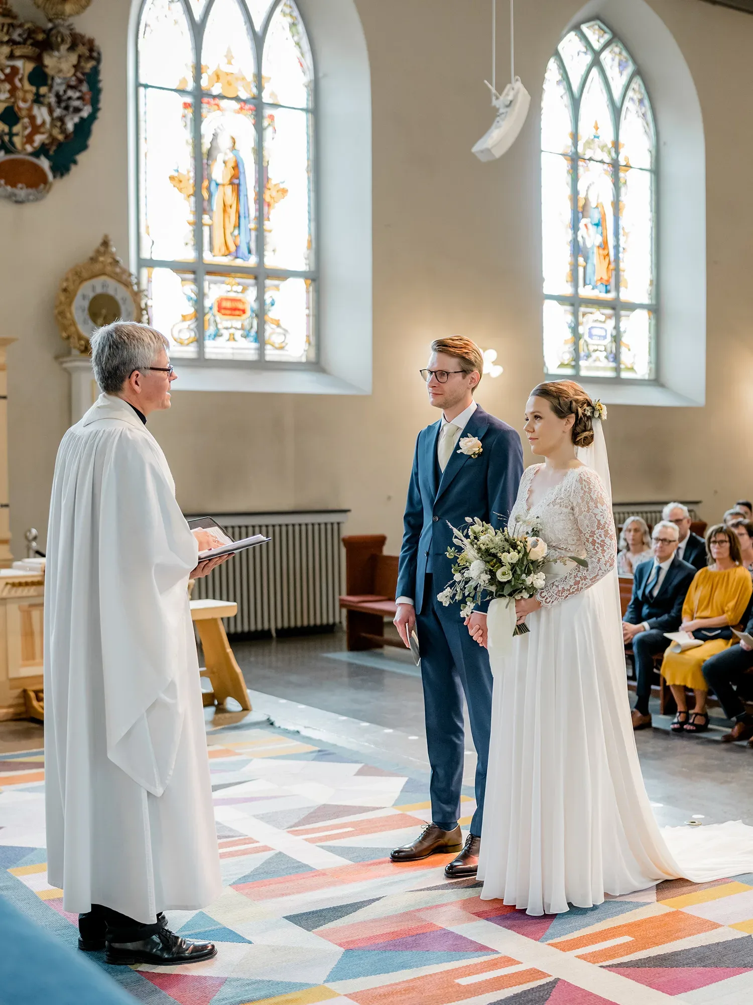 Das Brautpaar steht während der kirchlichen Trauung vor dem Priester. Die Braut trägt ein langes Kleid mit Spitzenärmeln und einen Blumenstrauß, der Bräutigam einen blauen Anzug. Hinter ihnen leuchten bunte Kirchenfenster.