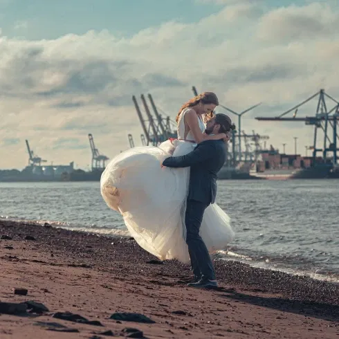 Ein Bräutigam hebt seine Braut am Ufer eines Flusses in die Luft. Im Hintergrund sind Hafenkräne und ein wolkiger Himmel zu sehen. Die Braut trägt ein voluminöses weißes Kleid.