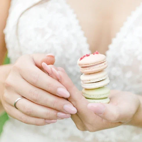 Nahaufnahme einer Braut, die eine kleine Stapelung von drei bunten Macarons in der Hand hält. Die Bräutigam trägt einen zarten Ring, und ihr Spitzenkleid ist im Hintergrund leicht erkennbar.