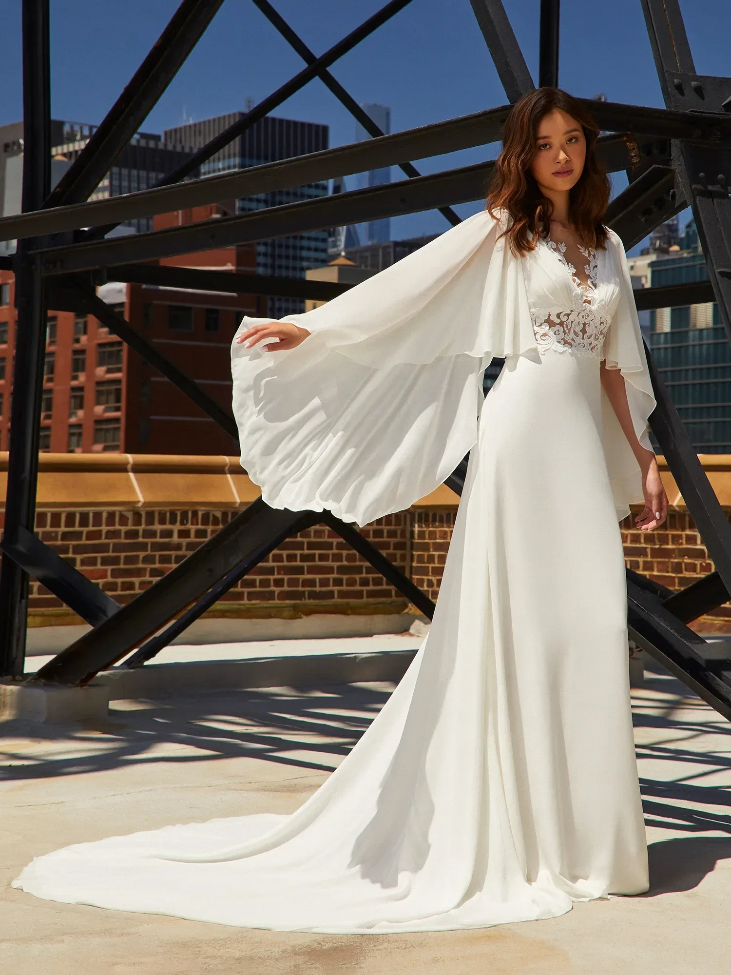 Eine Braut steht auf einer Dachterrasse und trägt ein fließendes, weißes Brautkleid mit Spitzenapplikationen und dramatischen, weit ausgestellten Ärmeln. Im Hintergrund sind moderne Gebäude zu sehen, die das urbane Ambiente betonen.