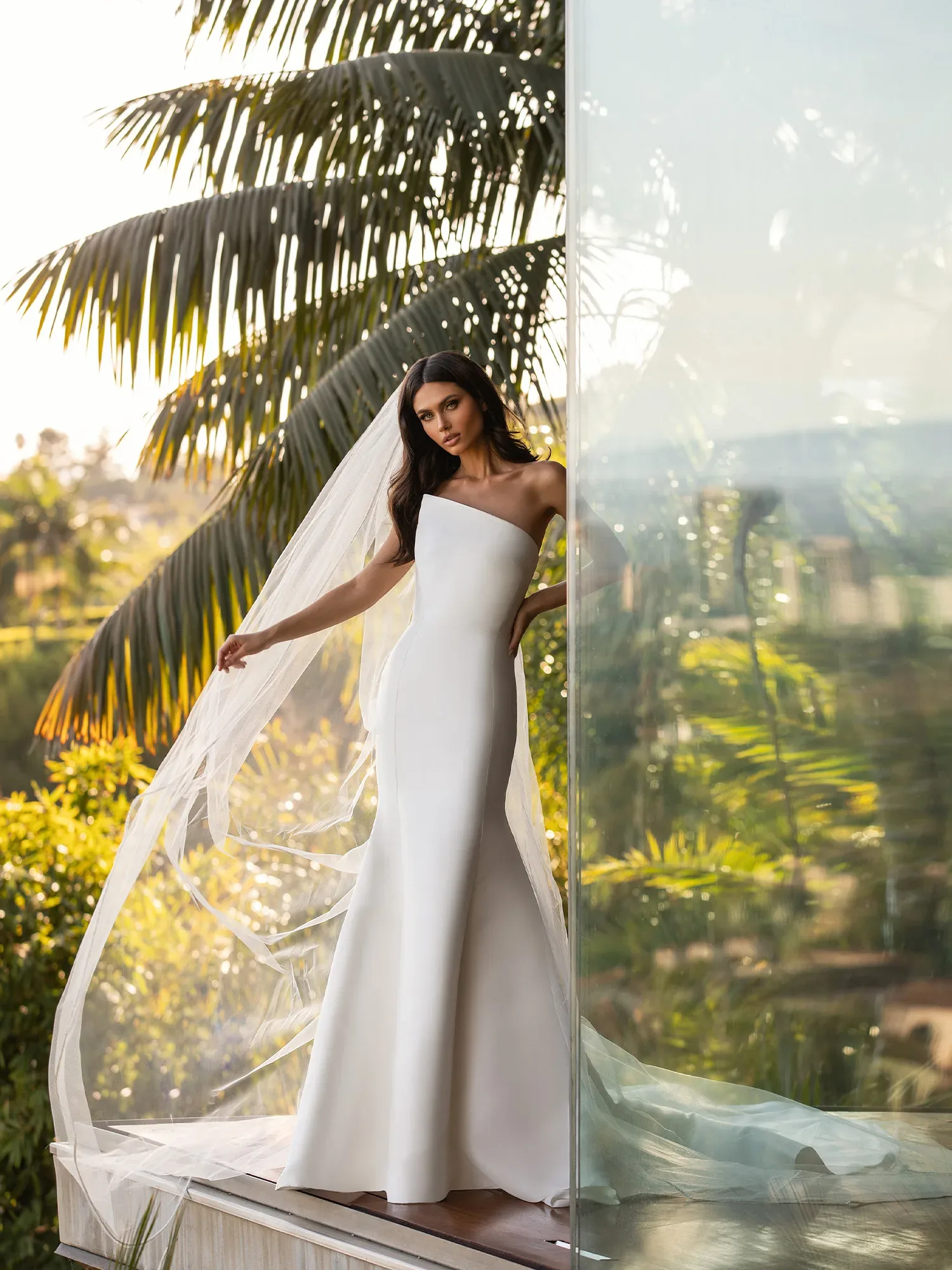 Eine elegante Braut steht in einem modernen, einseitig schulterfreien Brautkleid auf einer Terrasse vor üppigem Grün und Palmen. Ihr bodenlanger Schleier wird sanft vom Wind bewegt, während sie in die Ferne schaut.