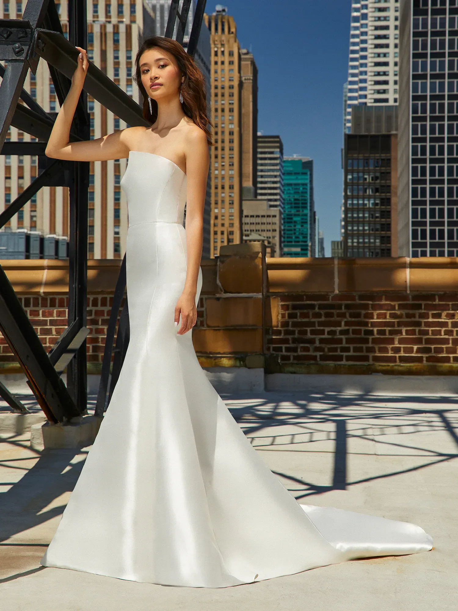 Eine schlanke Braut posiert in einem schulterfreien, eleganten Satin-Brautkleid auf einer städtischen Dachterrasse. Die klare Silhouette des Kleides betont ihre Figur, während die Hochhäuser im Hintergrund einen modernen urbanen Charme verleihen.