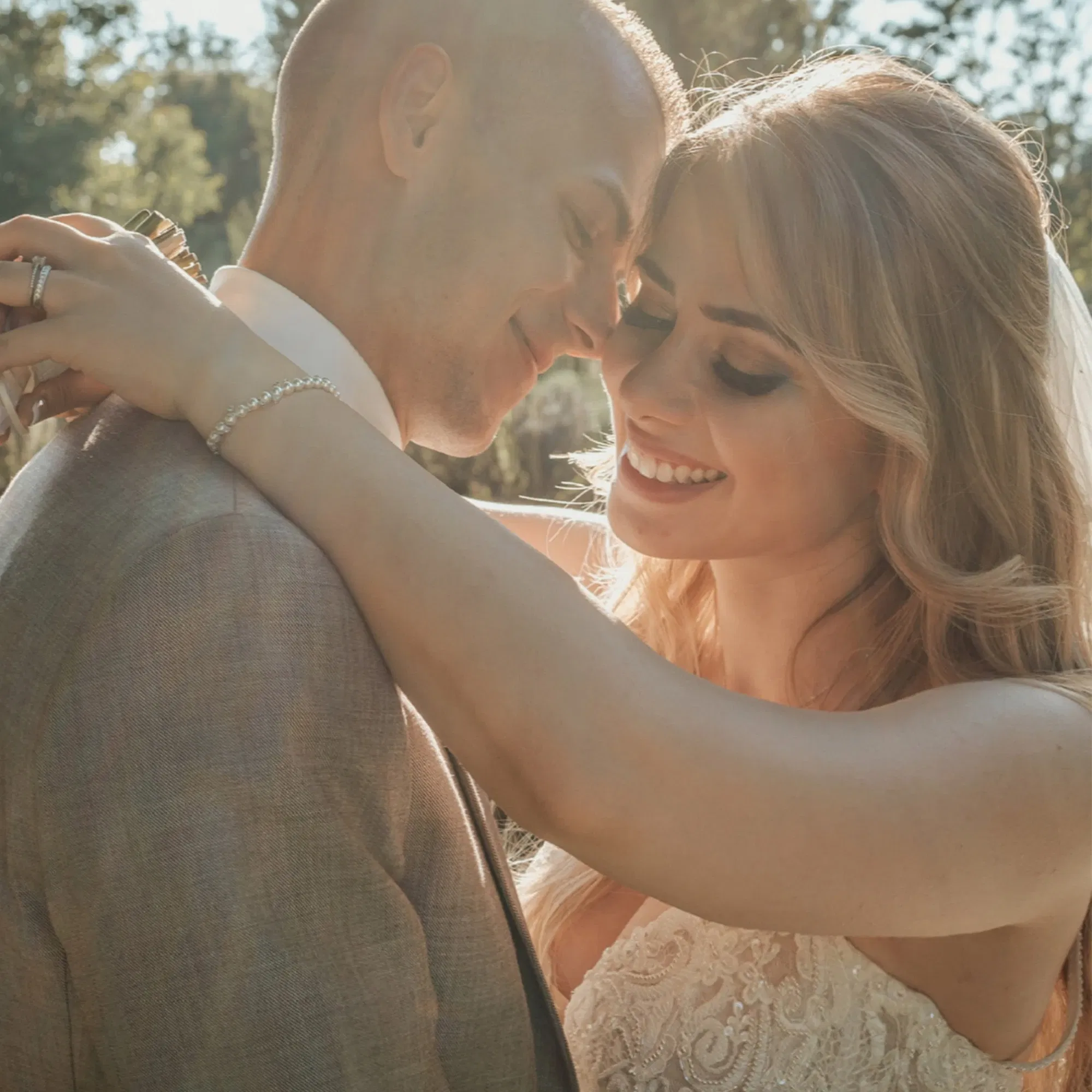 Nahaufnahme eines glücklichen Brautpaars, das sich lächelnd in die Arme schließt. Die Braut trägt ein Spitzenkleid und der Bräutigam einen grauen Anzug, während sie sich in einem Moment der Intimität im Freien genießen.