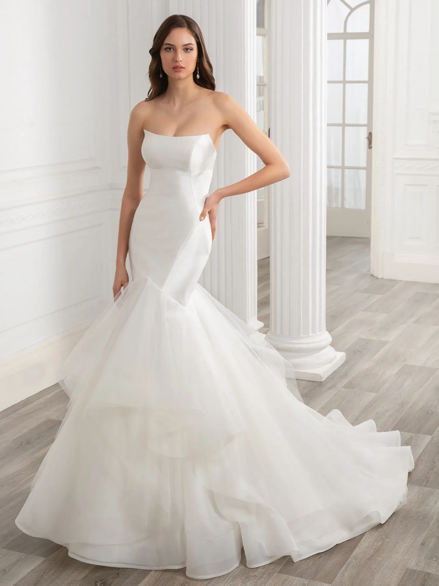 Eine elegante Braut trägt ein schulterfreies, figurbetontes Brautkleid mit fließendem Tüllrock. Sie steht vor einer weißen, klassischen Säulen-Architektur und blickt gerade in die Kamera.