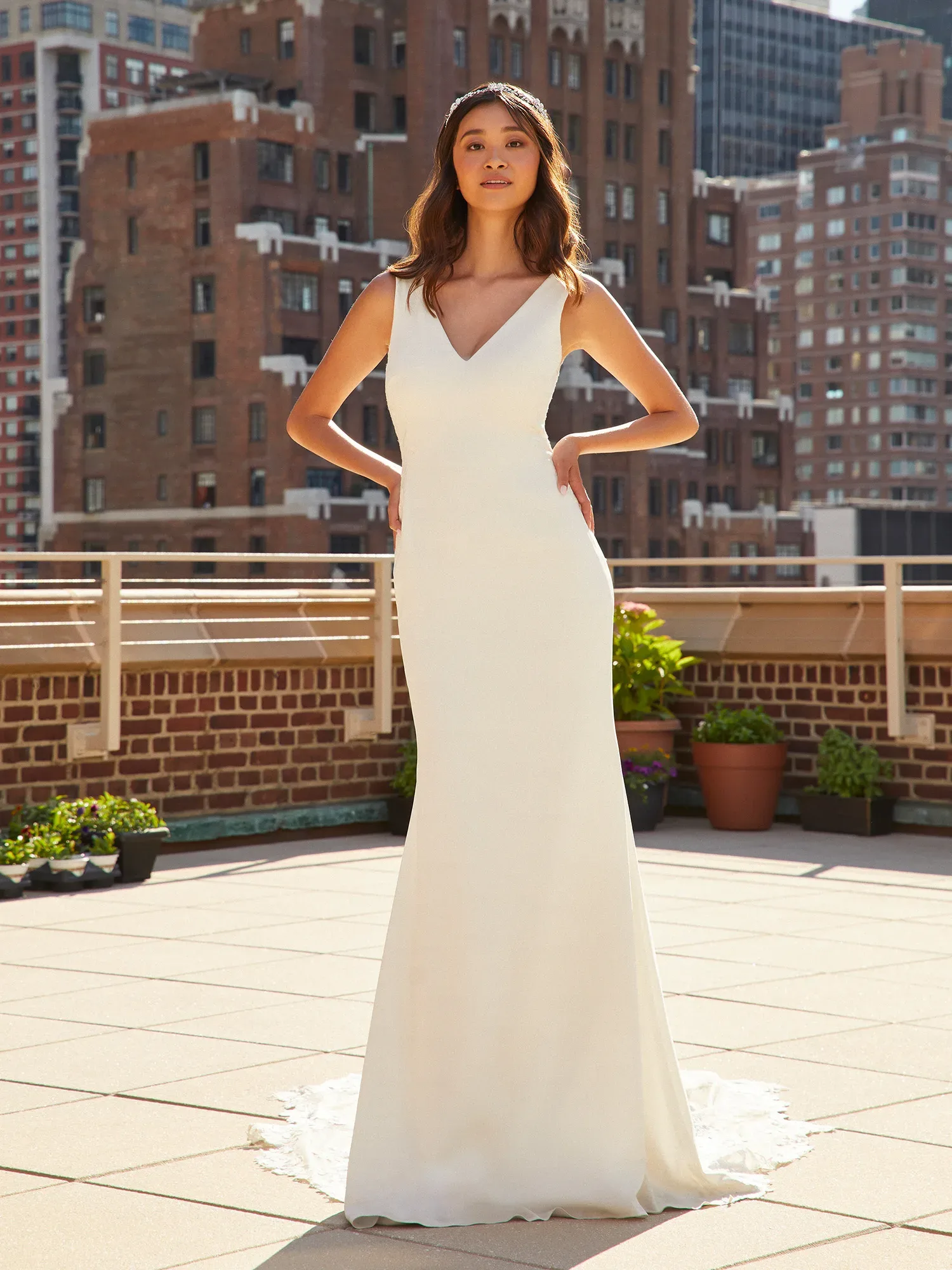 Eine Frau steht in einem eleganten, schlichten Brautkleid mit V-Ausschnitt auf einer städtischen Dachterrasse. Im Hintergrund sind hohe Backsteingebäude zu sehen, und die Braut posiert selbstbewusst mit den Händen an der Hüfte.