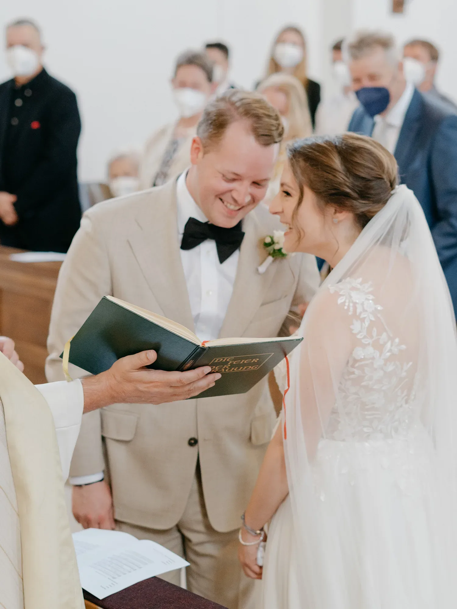 Das Brautpaar lacht während der kirchlichen Zeremonie, während der Priester aus einem Buch vorliest. Beide stehen Seite an Seite, und der Bräutigam trägt einen beigen Anzug mit schwarzer Fliege. Die Gäste im Hintergrund tragen Masken.